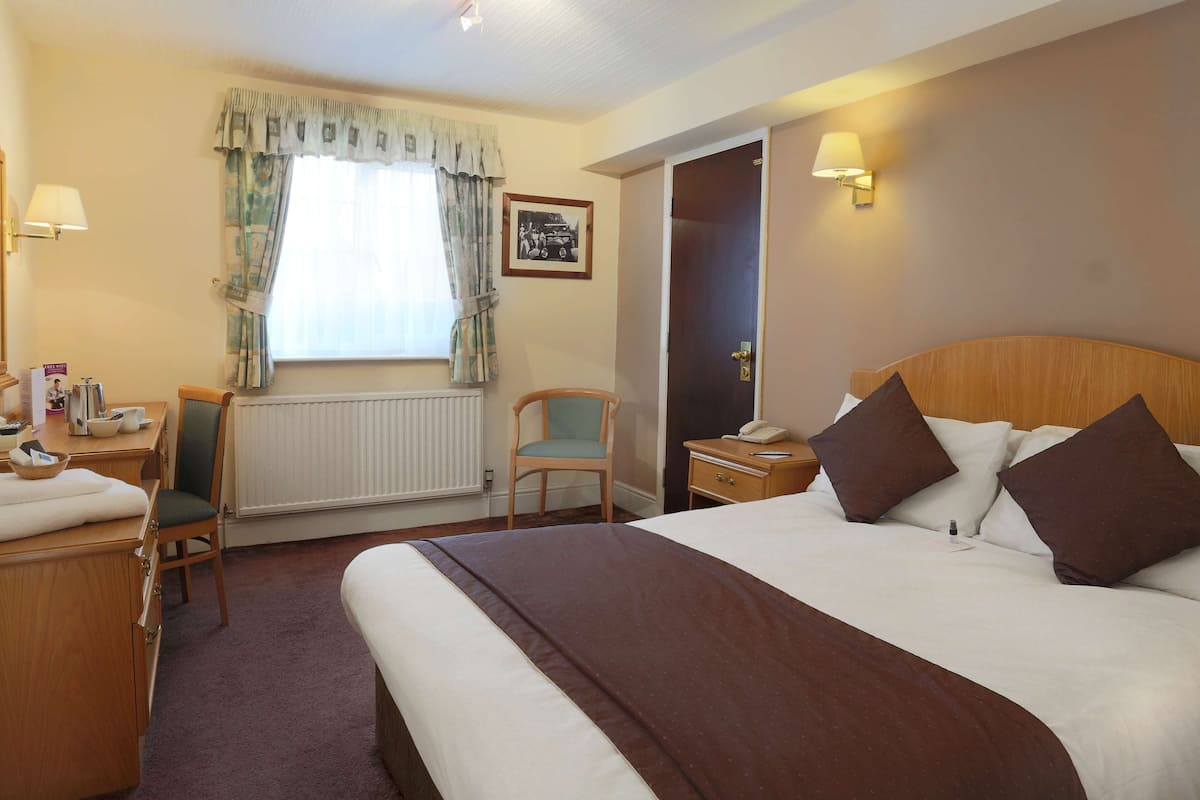 A hotel room with a large bed, a desk with a chair, a bedside table with a lamp, and a window with curtains.