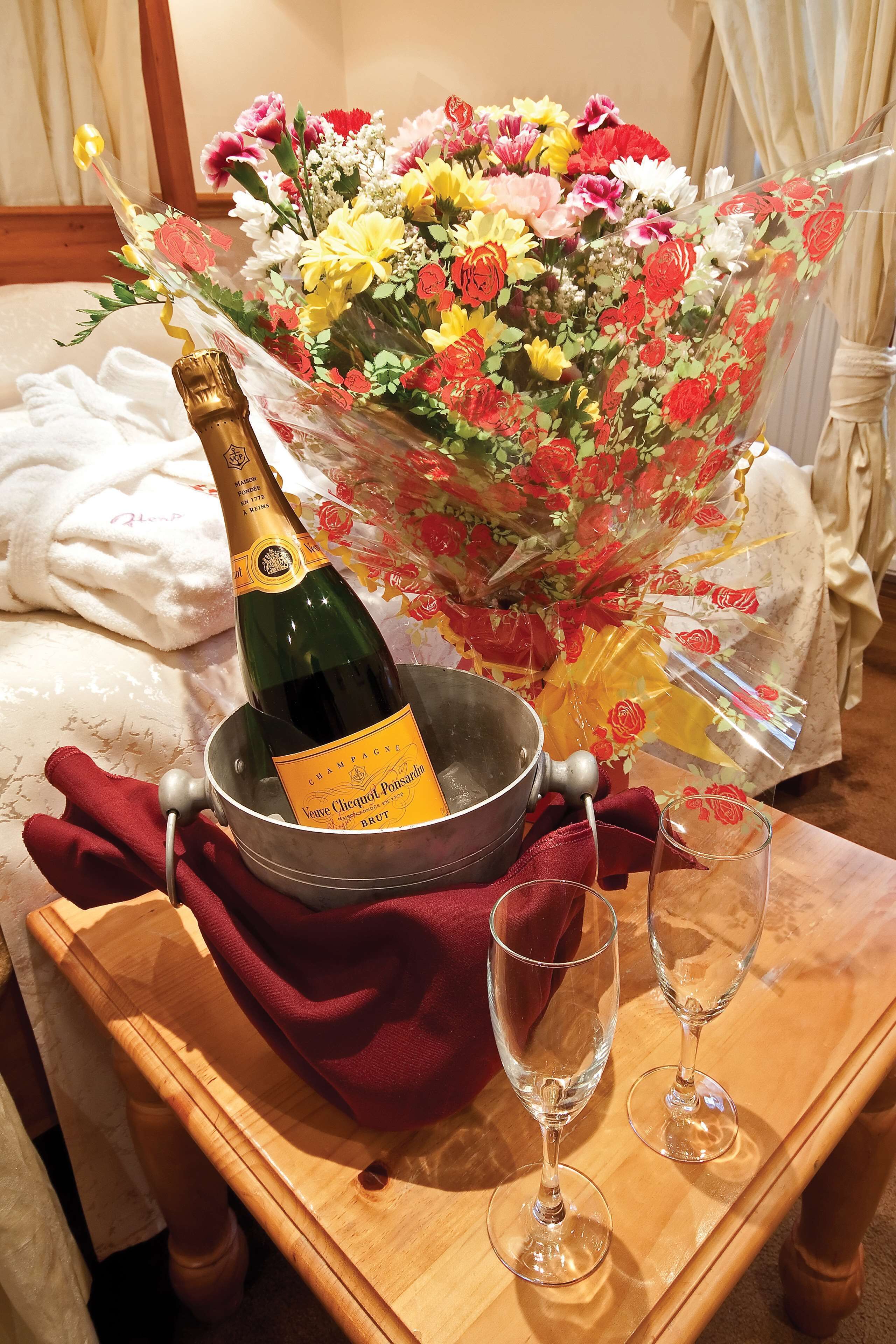A bottle of champagne in a bucket, a bouquet of flowers, and two wine glasses on a wooden table.