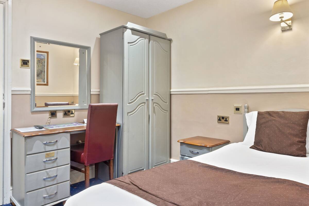 A hotel room with a bed, a desk with drawers, a chair, a wardrobe, and a nightstand.
