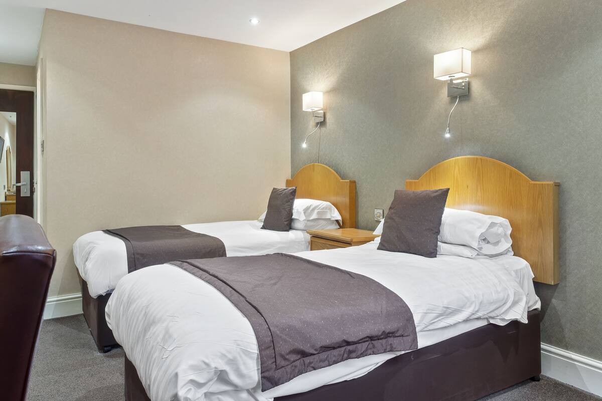 A hotel room with two beds, each with a wooden headboard, white bedding, and a brown accent pillow. There are wall-mounted nightstands with lamps, and a door leading to another room.
