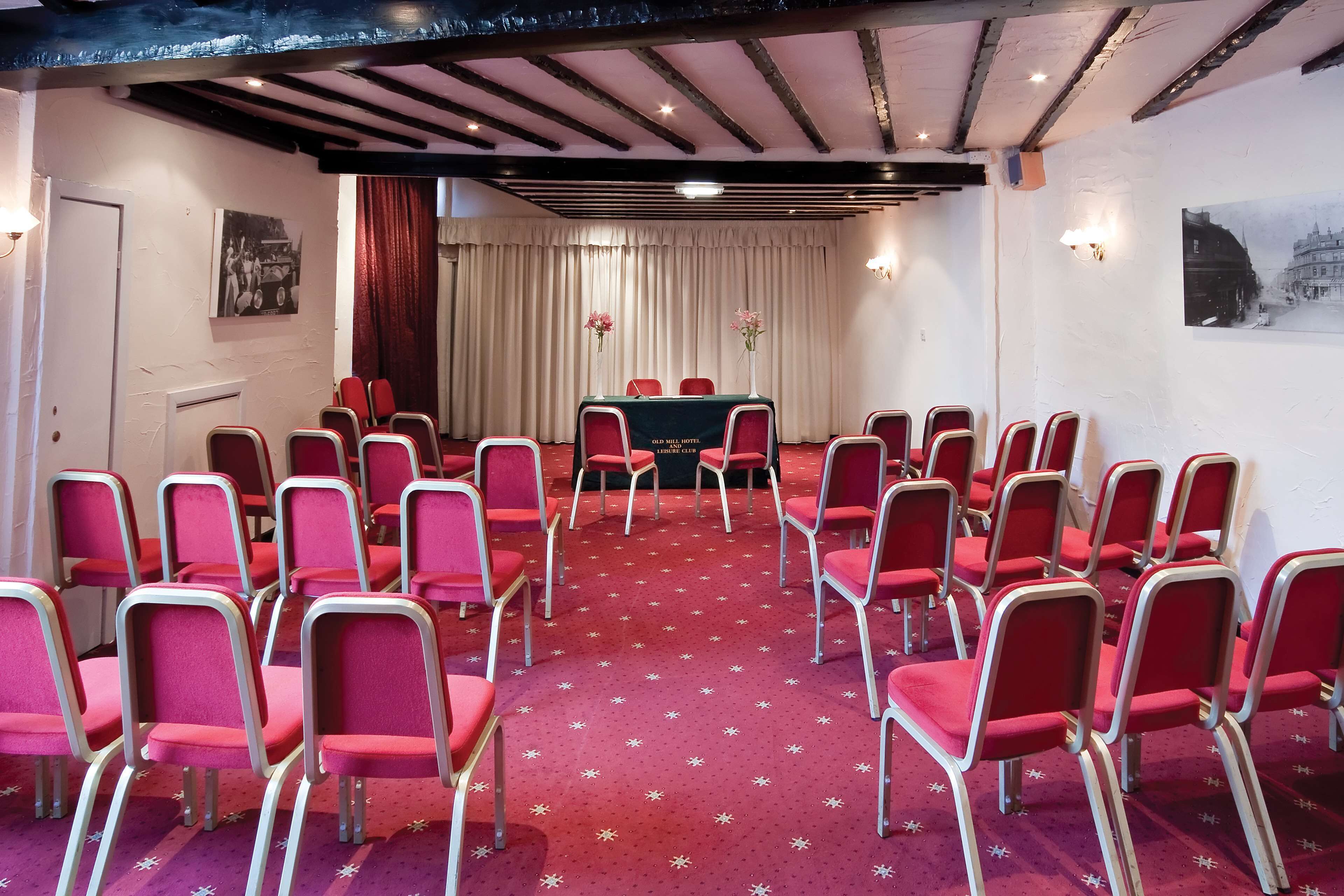 Un salón de conferencias con sillas rojas dispuestas en filas, una mesa con mantel verde y una alfombra roja con motivos de estrellas.