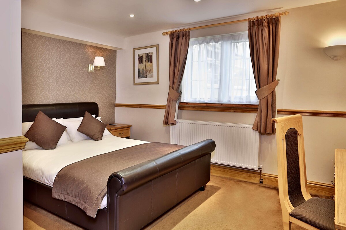 A hotel room with a bed, a chair, a nightstand, a lamp, a window with curtains, and a radiator.