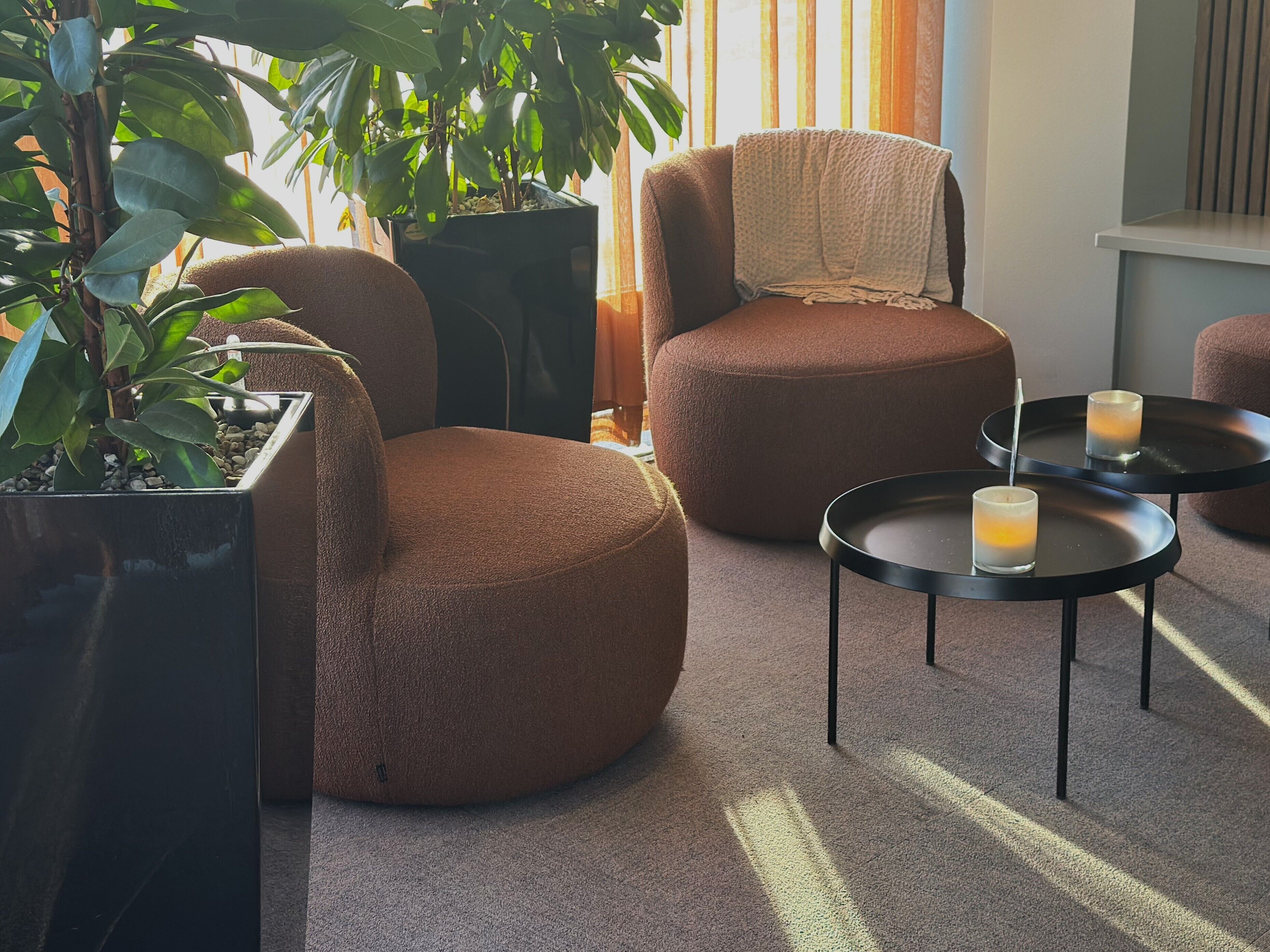 Un coin salon accueillant avec deux fauteuils ronds de couleur marron et une petite table sur laquelle sont posées des bougies. On y trouve également des plantes en pot et une grande fenêtre ornée de rideaux.
