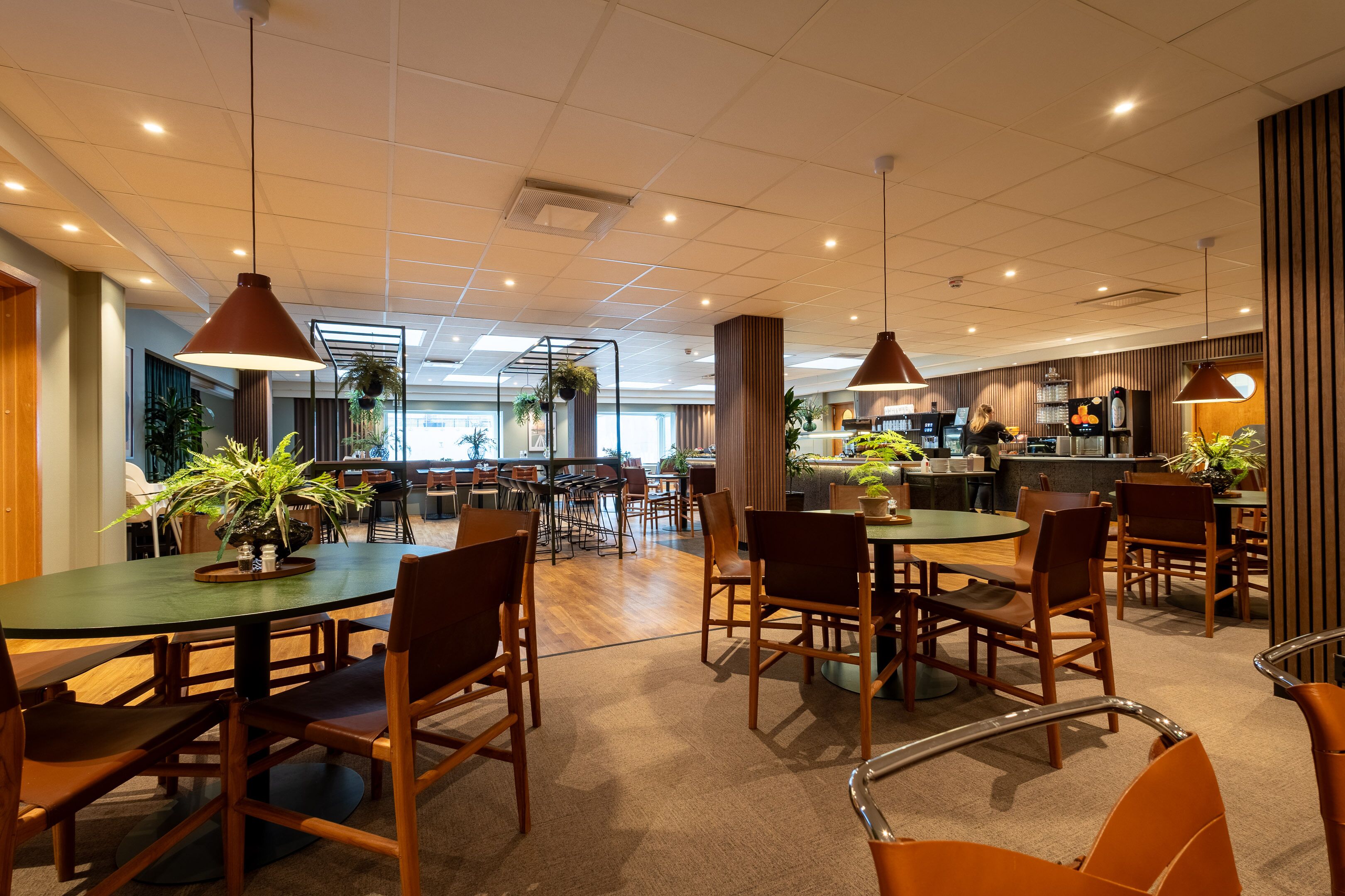 A spacious indoor dining area with multiple tables and chairs, a kitchen in the background, and potted plants.