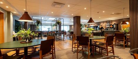 A spacious indoor dining area with multiple tables and chairs, a kitchen in the background, and potted plants.