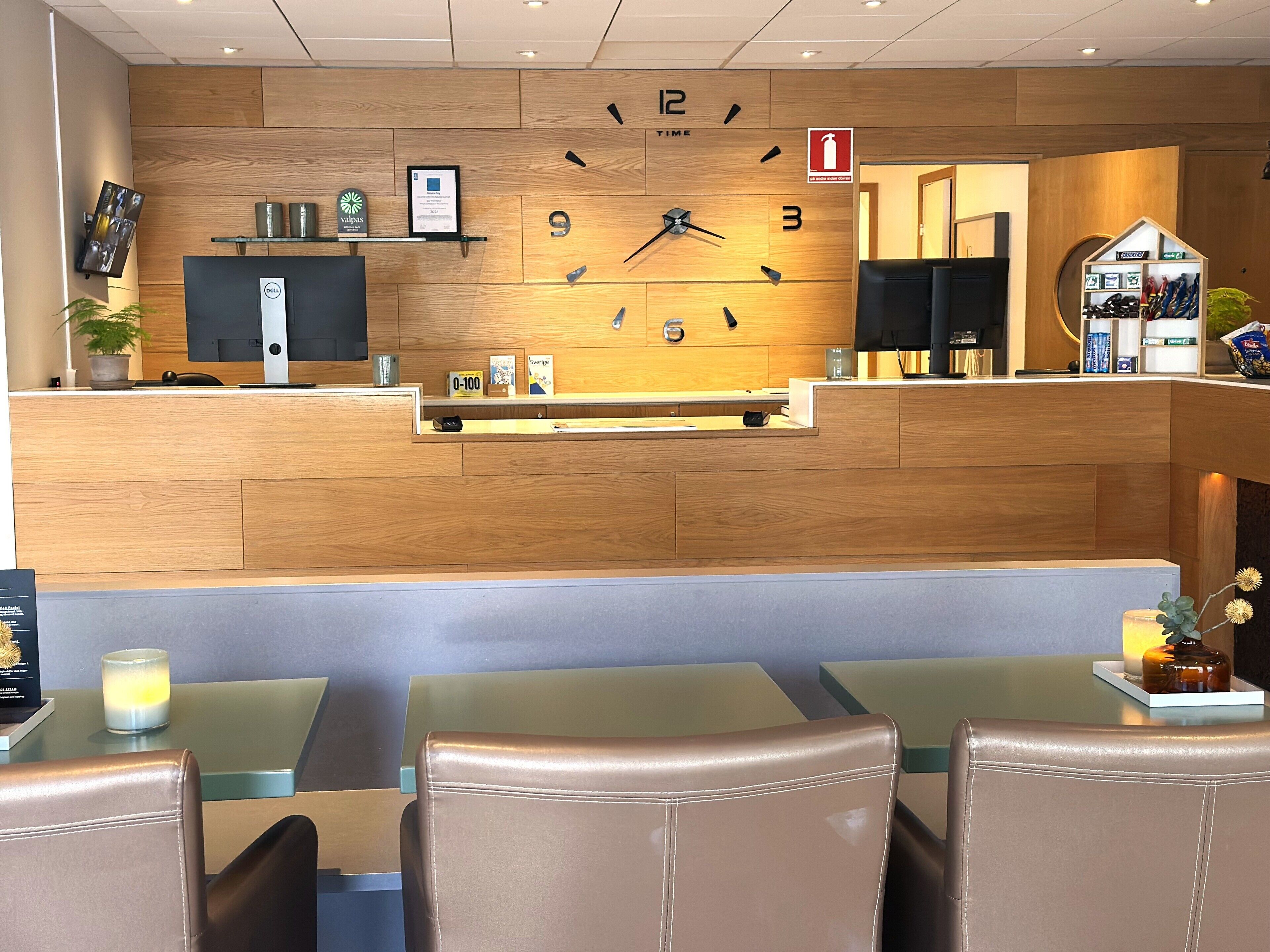 A hotel reception area with a wooden reception desk, a wall clock, and seating area with tables and chairs.