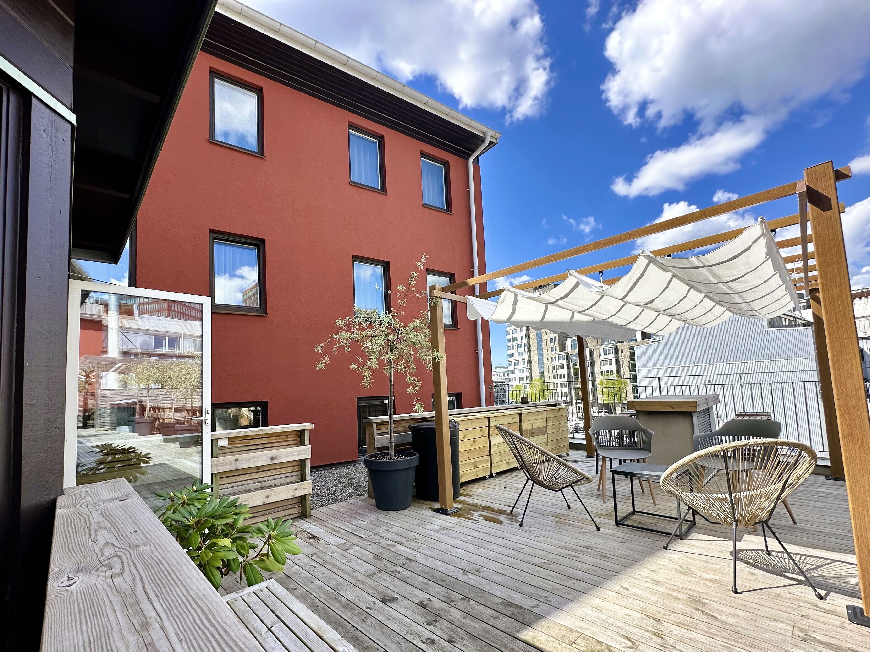 En träaltan med utemöbler, en pergola med ett vitt tak och en röd byggnad i bakgrunden.