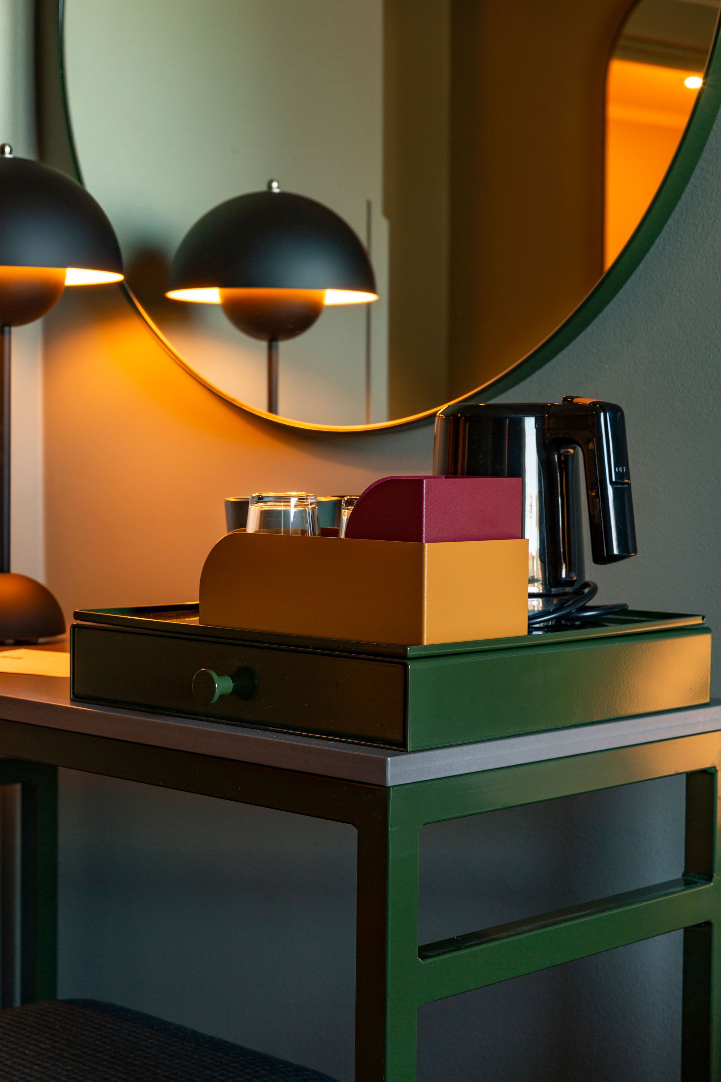 A desk with a mirror, a lamp, a coffee maker, and a box.