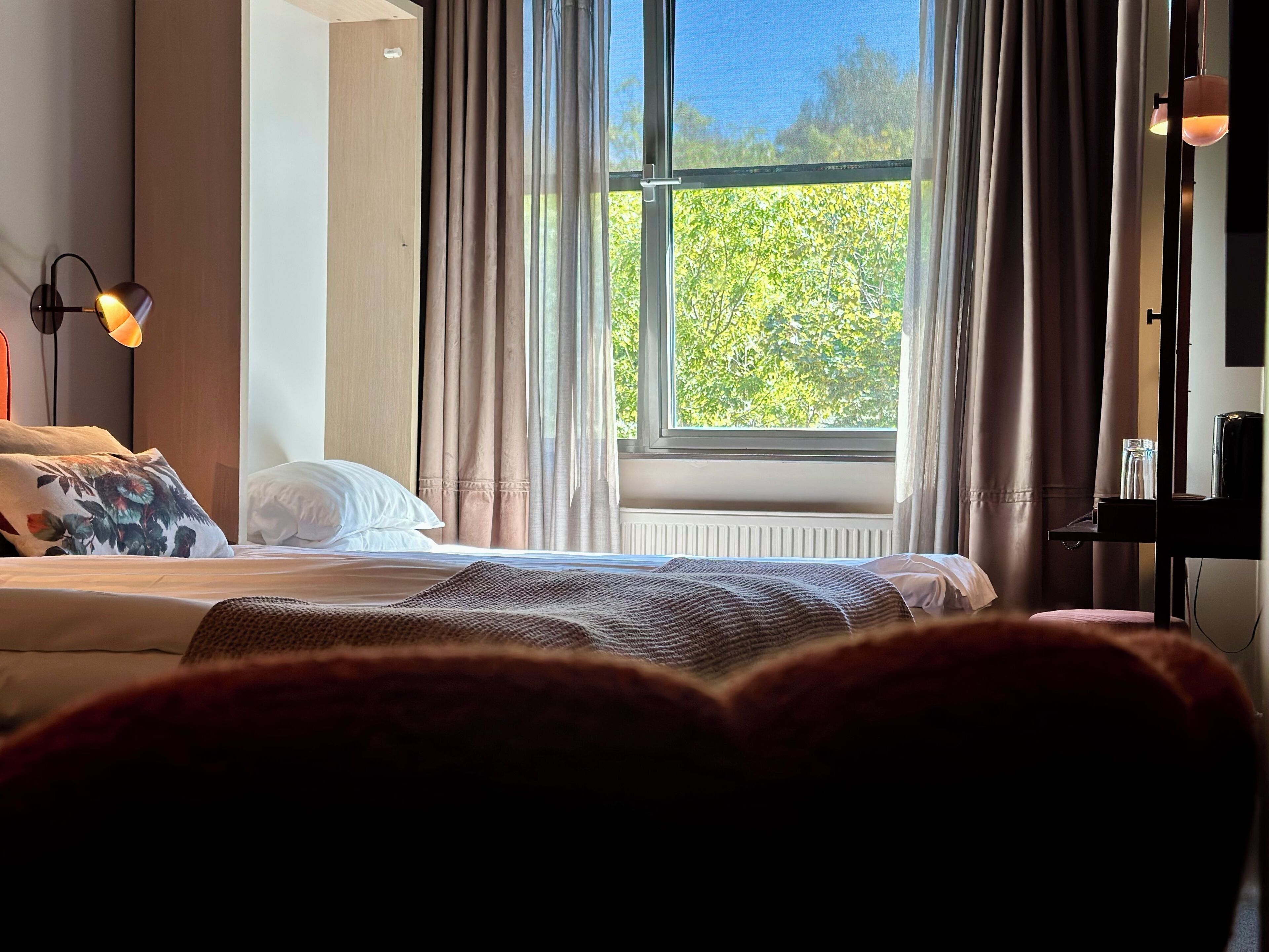 Hotellihuone, jossa on sänky, ikkuna verhoilla, lamppu ja näkymä vihreälle alueelle.