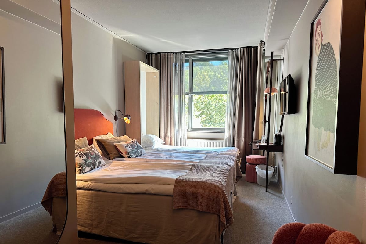 Una habitació d'hotel amb un llit gran, una taula de nit, una cadira, una finestra amb cortines i un televisor fixat a la paret.