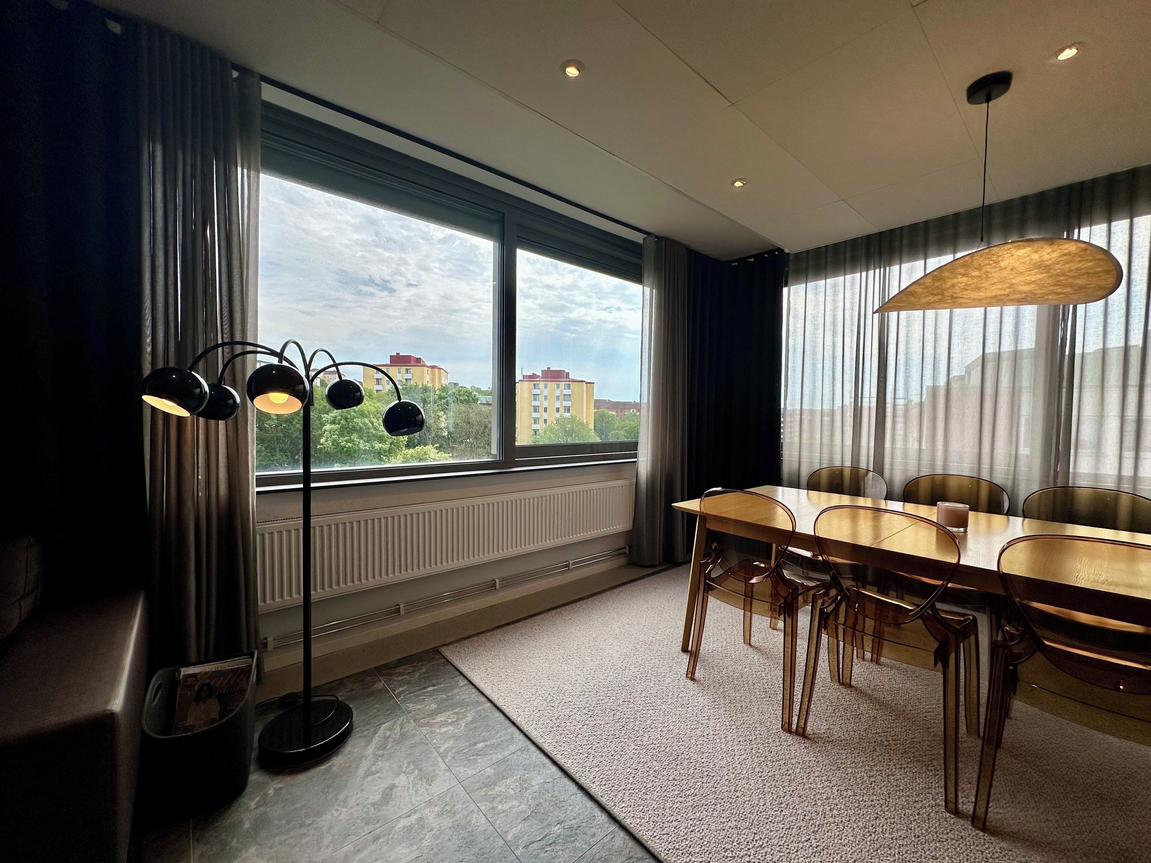 Ein moderner Essbereich mit großem Tisch, Stühlen, einer Stehlampe und Blick auf Gebäude durch große Fenster.