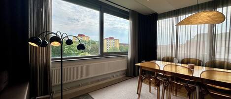 A modern dining area with a large table, chairs, a floor lamp, and a view of buildings through large windows.