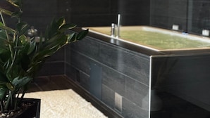 A modern bathroom with a dark tiled wall, a bathtub, a potted plant, and a woven rug.