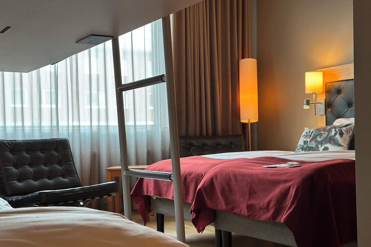 Habitació d'hotel amb un llit, una cadira, una taula de nit, una làmpada i vista per la finestra.