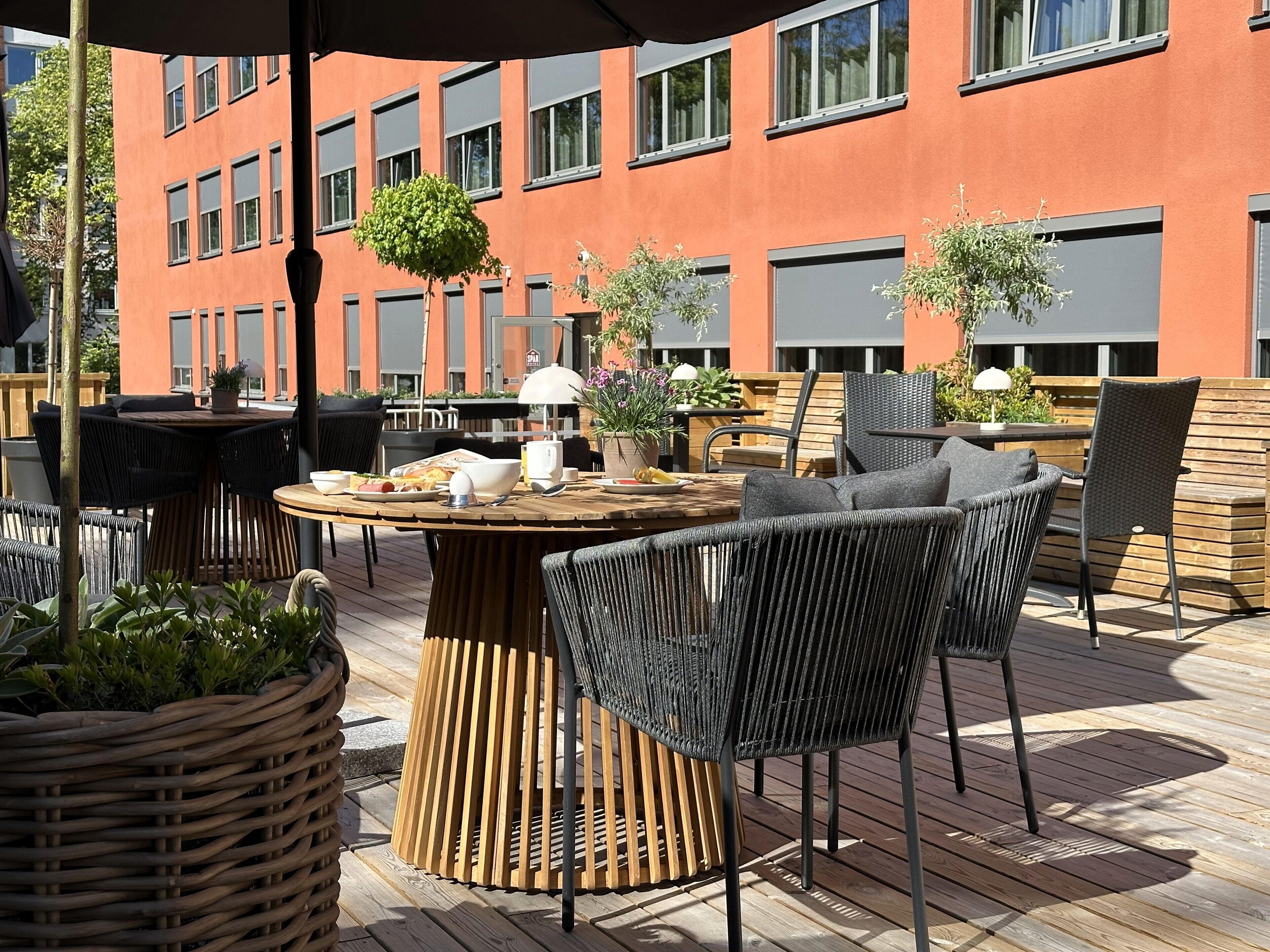 An outdoor patio with round tables and wicker chairs, surrounded by potted plants and a wooden deck.