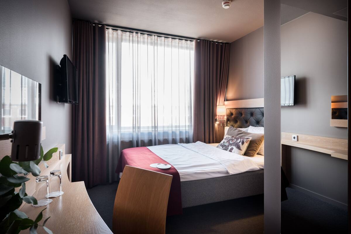 Habitació d'hotel amb llit, escriptori, cadira, televisió i finestra amb cortines.