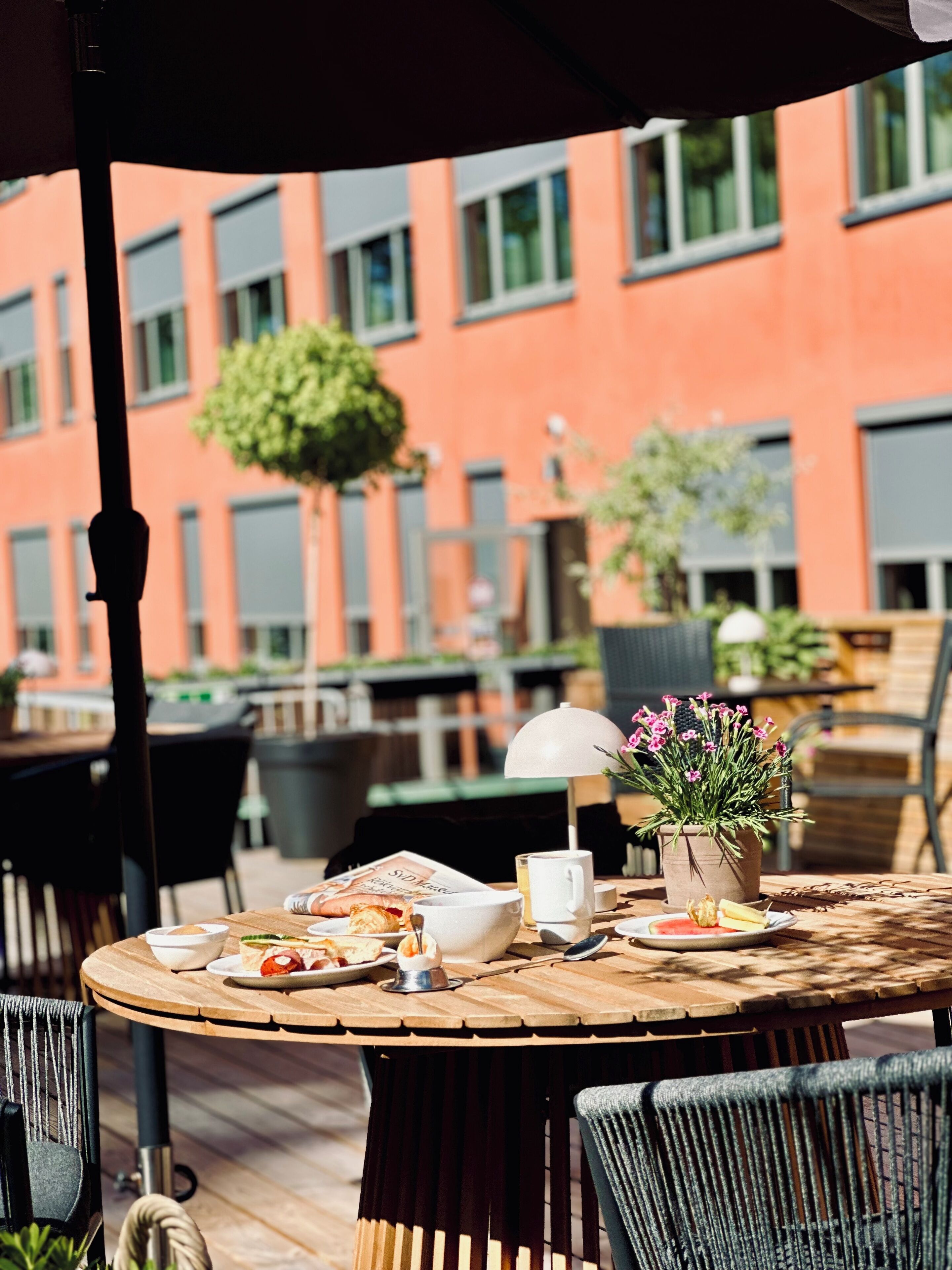 An outdoor dining area with a round wooden table, chairs, and a potted plant with pink flowers.