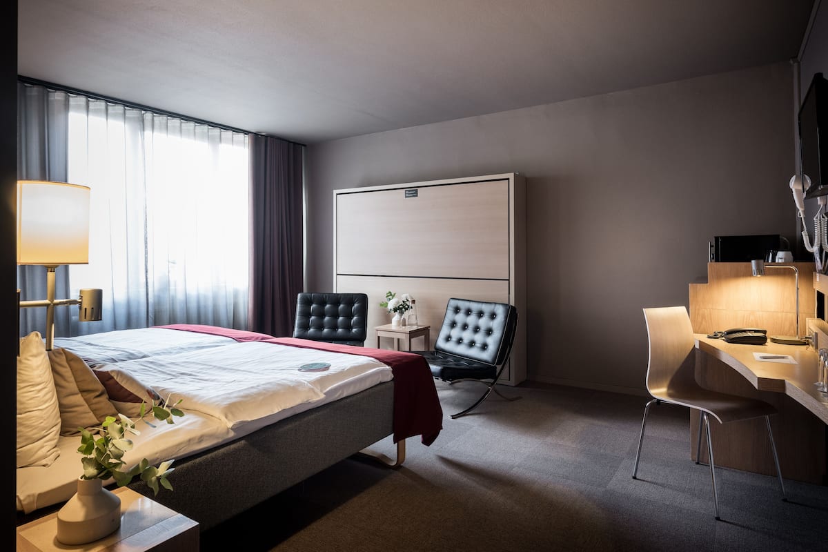 Habitació d'hotel amb un llit, una taula de treball, una cadira, un televisor i una finestra amb cortines.