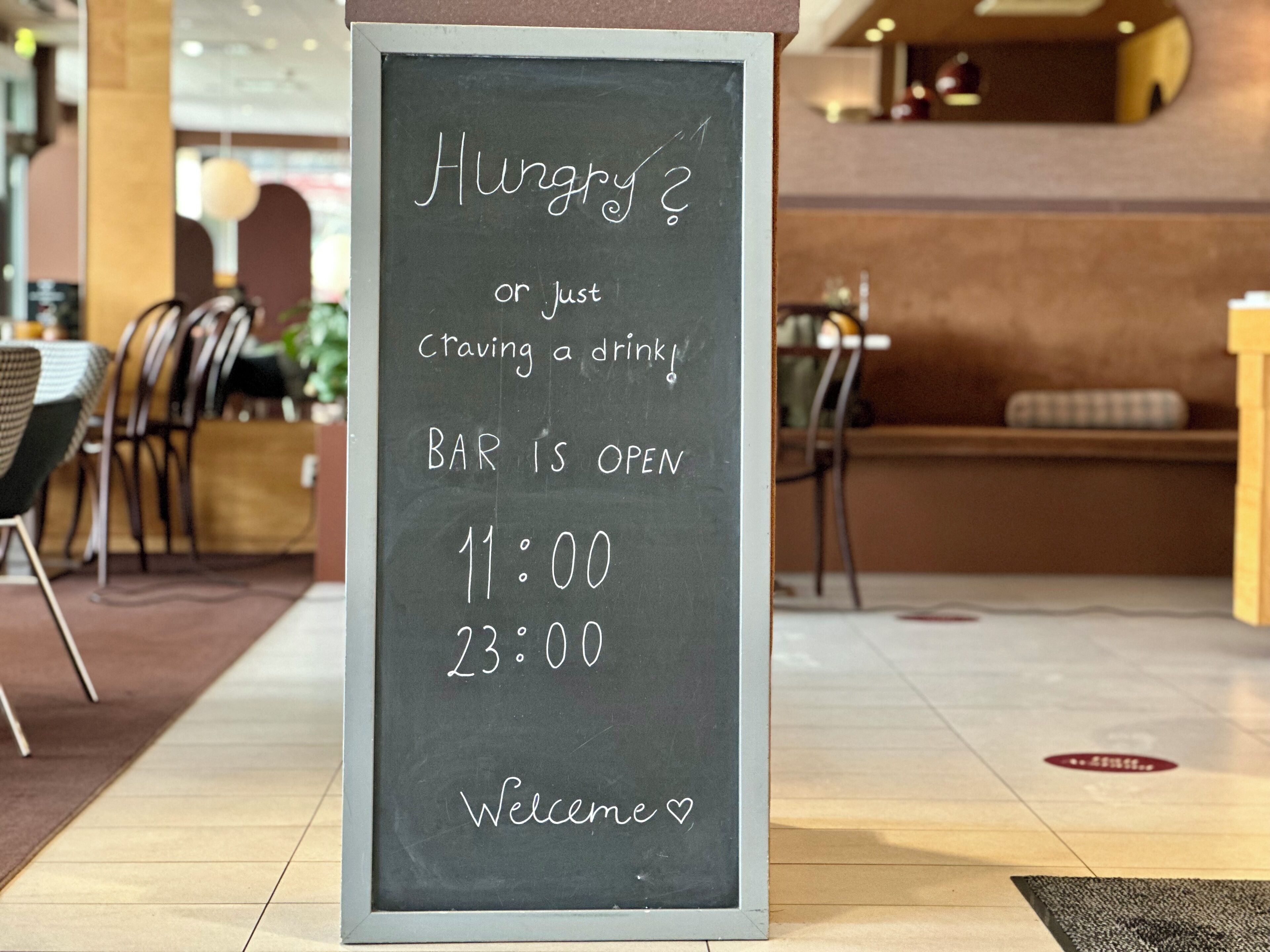 Una pizarra en una cafetería con el texto '¿Tienes hambre? o ¿solo te apetece algo de beber? BAR ABIERTO 11:00 23:00 ¡Bienvenidos!'