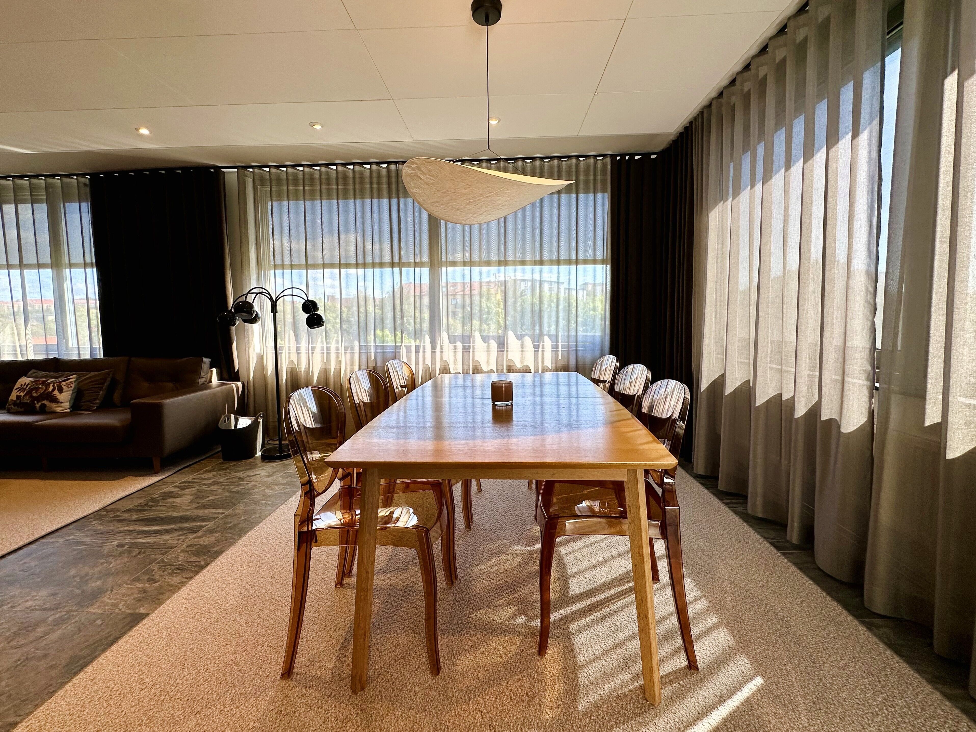 A modern meeting room with a long wooden table, multiple chairs, a sofa, and large windows with sheer curtains.