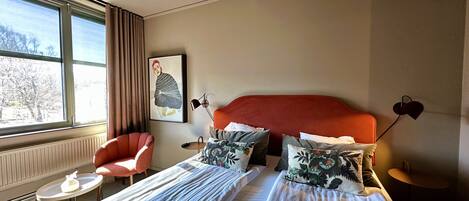 A hotel room with a large bed, a chair, a small table, and a window with a view of trees.