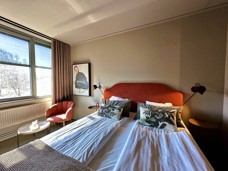 A hotel room with a large bed, a chair, a small table, and a window with a view of trees.
