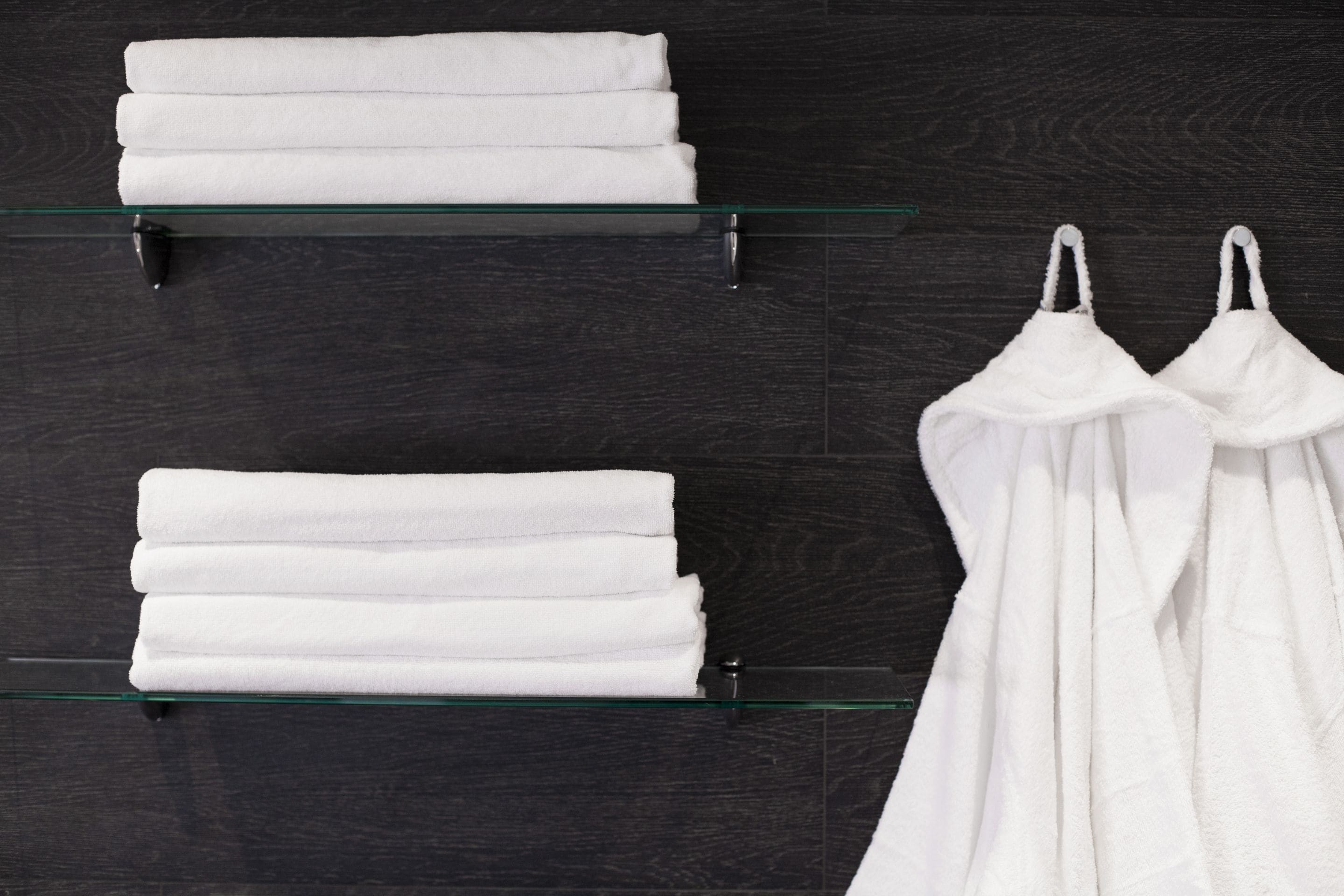 Un baño con dos repisas de vidrio que sostienen toallas blancas dobladas y un par de batas de baño blancas.