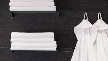 Un baño con dos repisas de vidrio que sostienen toallas blancas dobladas y un par de batas de baño blancas.