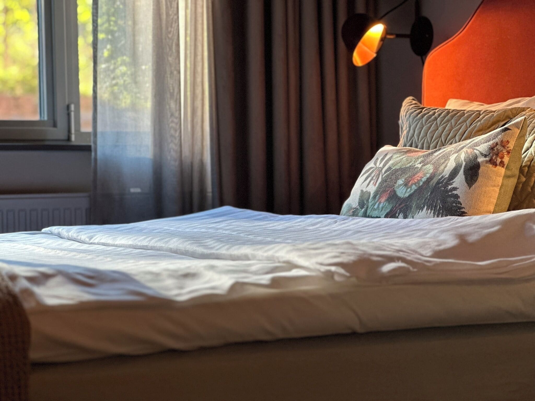 A bed with white bedding, a headboard with a floral pillow, and a lamp with an orange shade.