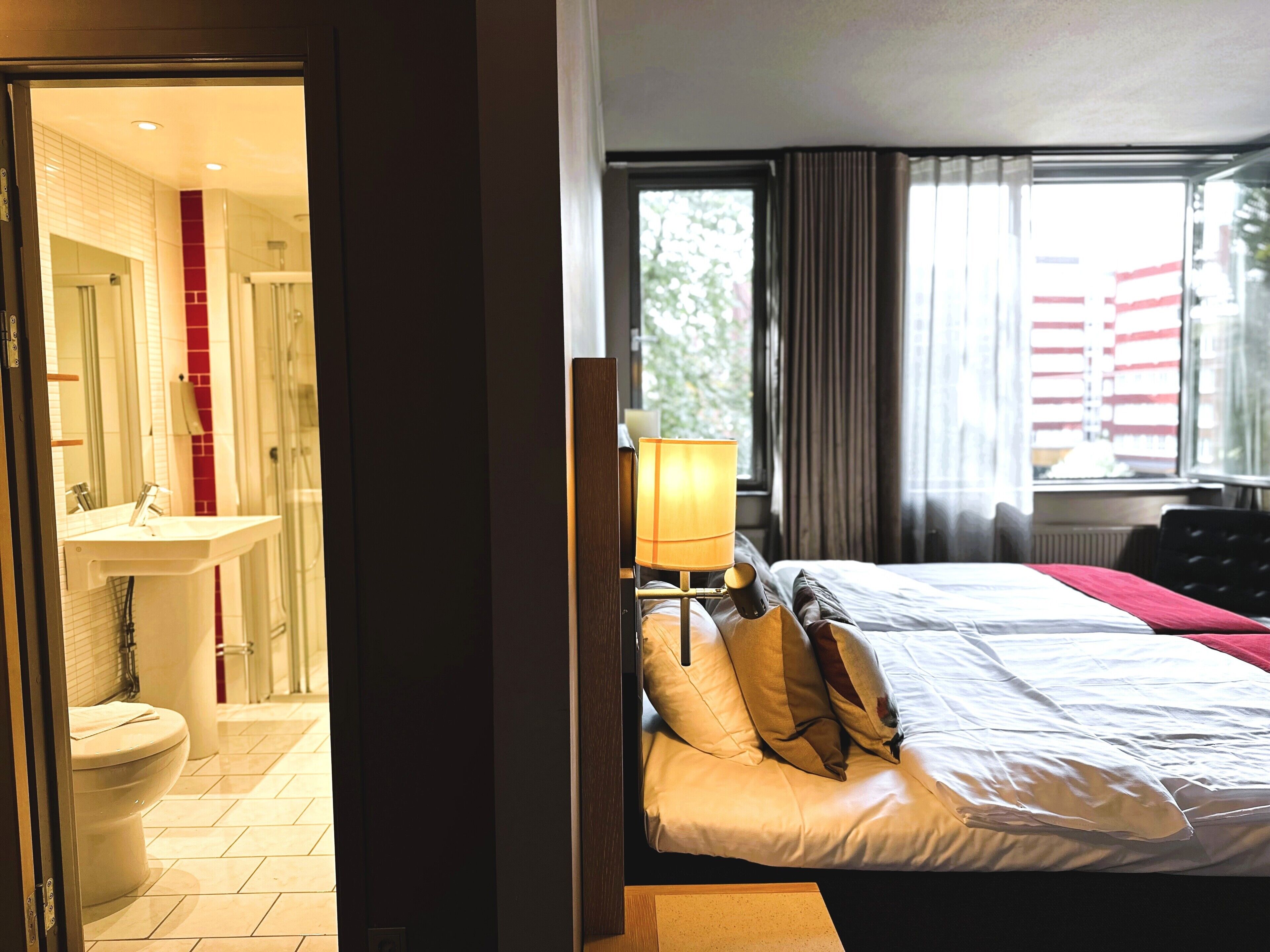 Ett hotellrum med en säng, en sänglampa, ett fönster med gardiner och ett badrum med handfat och toalett.