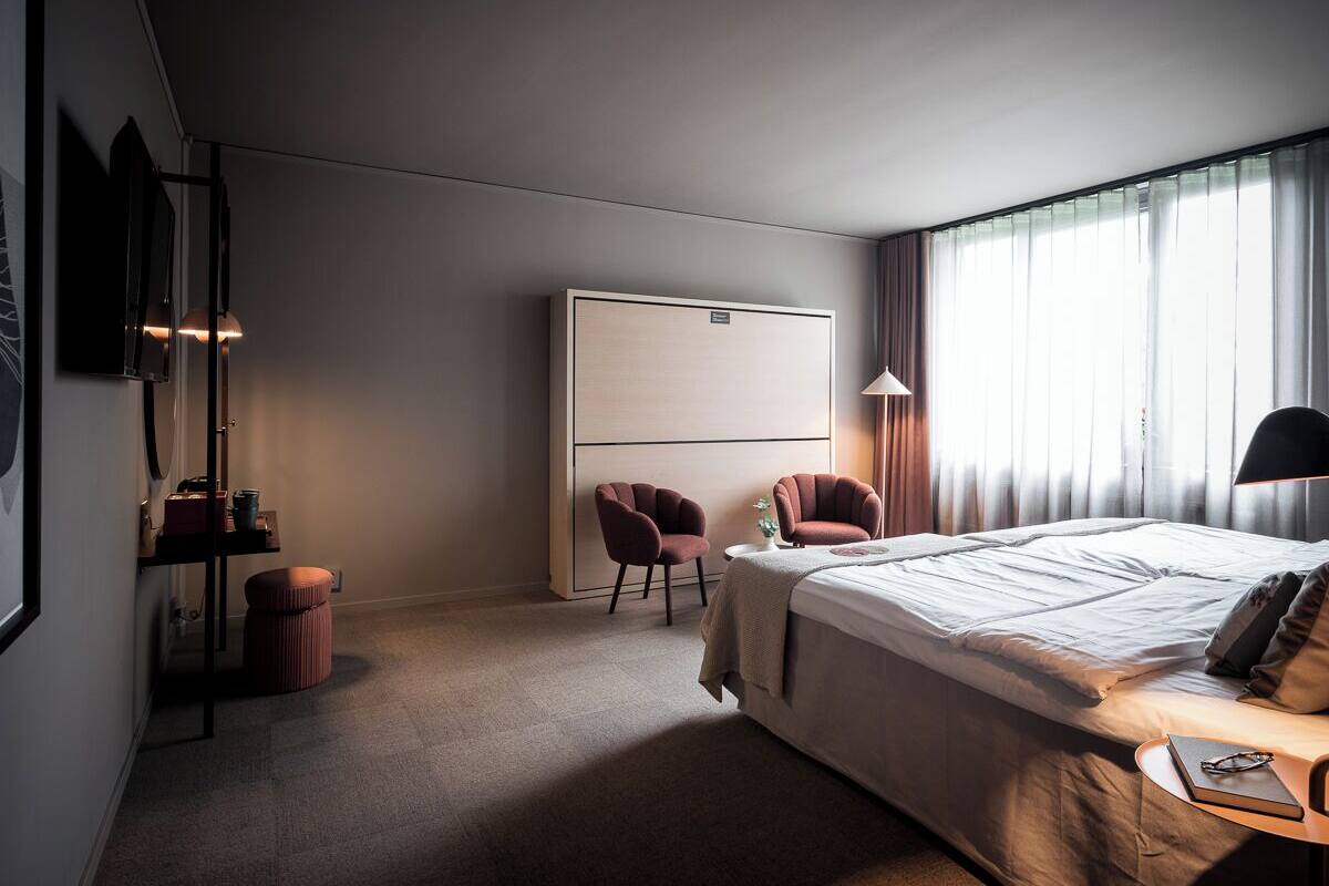 Habitació d'hotel amb un llit, dues cadires, un escriptori amb una làmpada, un televisor i una finestra gran amb cortines.