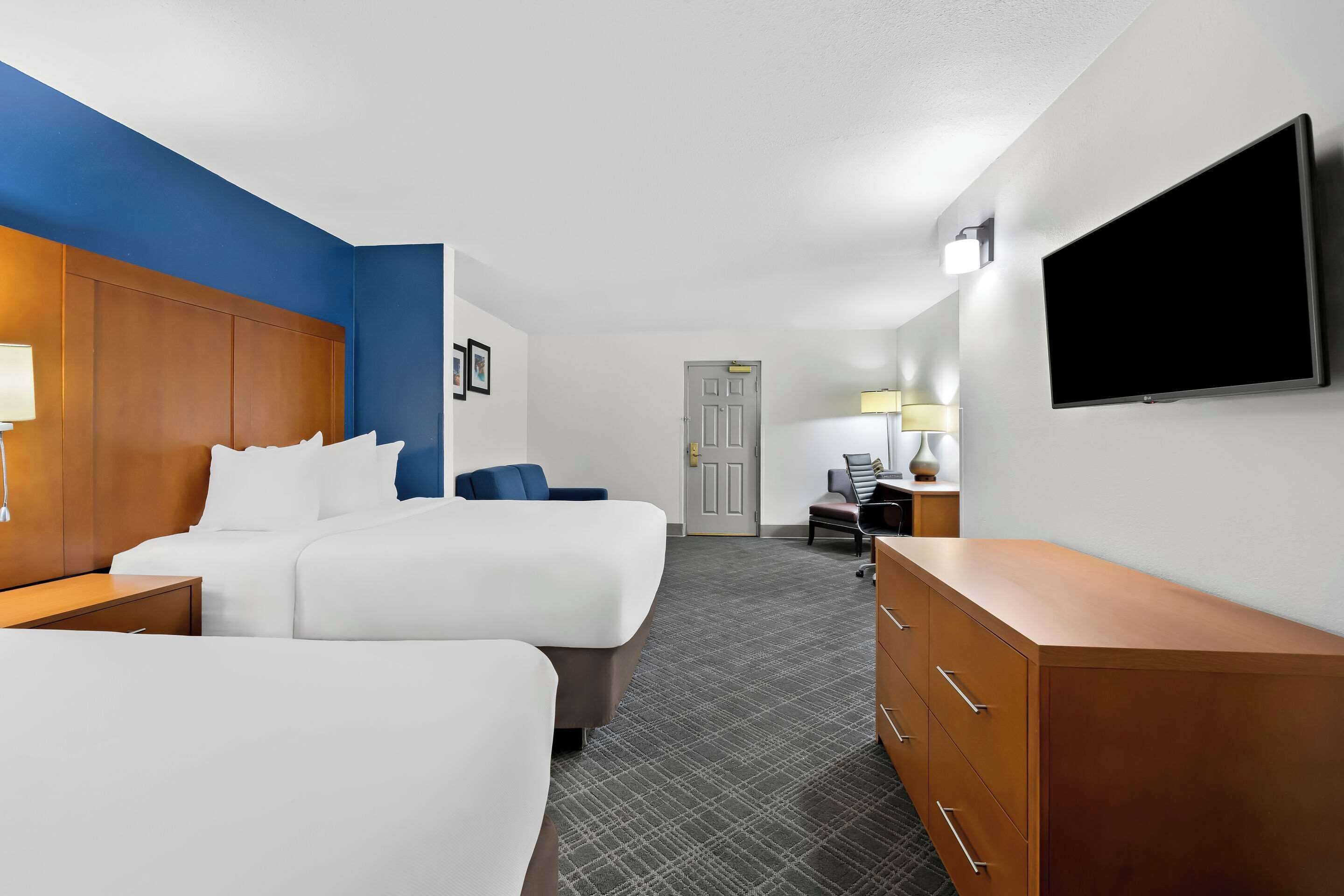 Habitación de hotel con dos camas, un escritorio, un aparador, un televisor y una puerta que da a otra habitación.