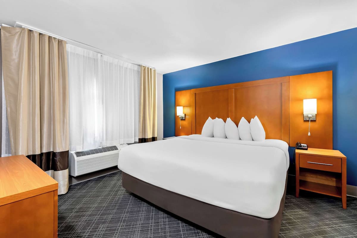 A hotel room with a large bed, a bedside table, a nightstand, a wall-mounted lamp, and a window with curtains.