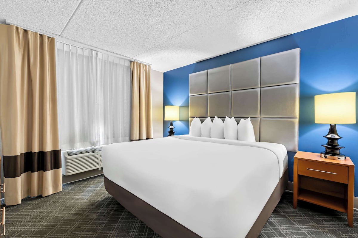 A hotel room with a large bed, a headboard with multiple panels, two bedside lamps, a heater, and a window with curtains.