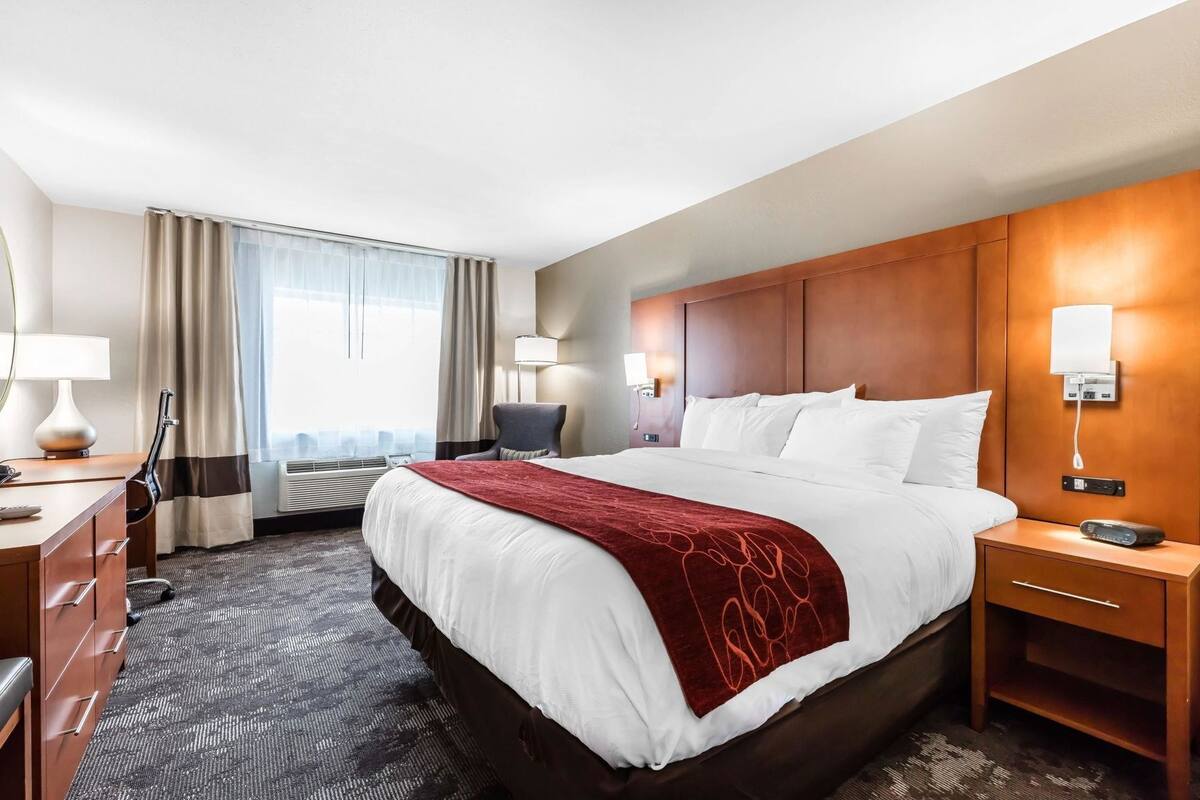 A hotel room with a large bed, a desk with a lamp, a chair, a bedside table with a lamp, and a view of the city through the window.