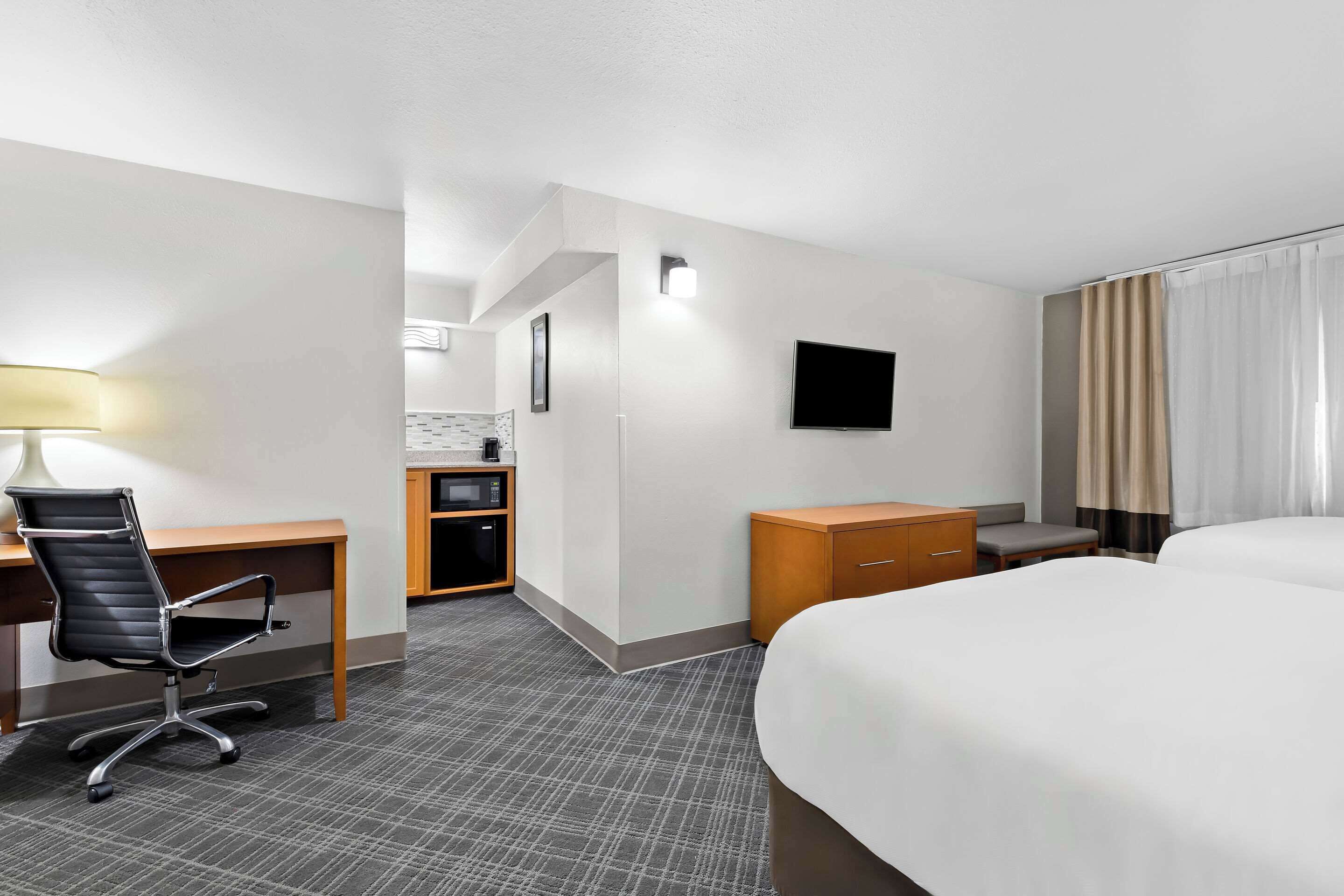 A hotel room with a bed, a desk with a chair, a TV, a small kitchenette, and a bathroom.