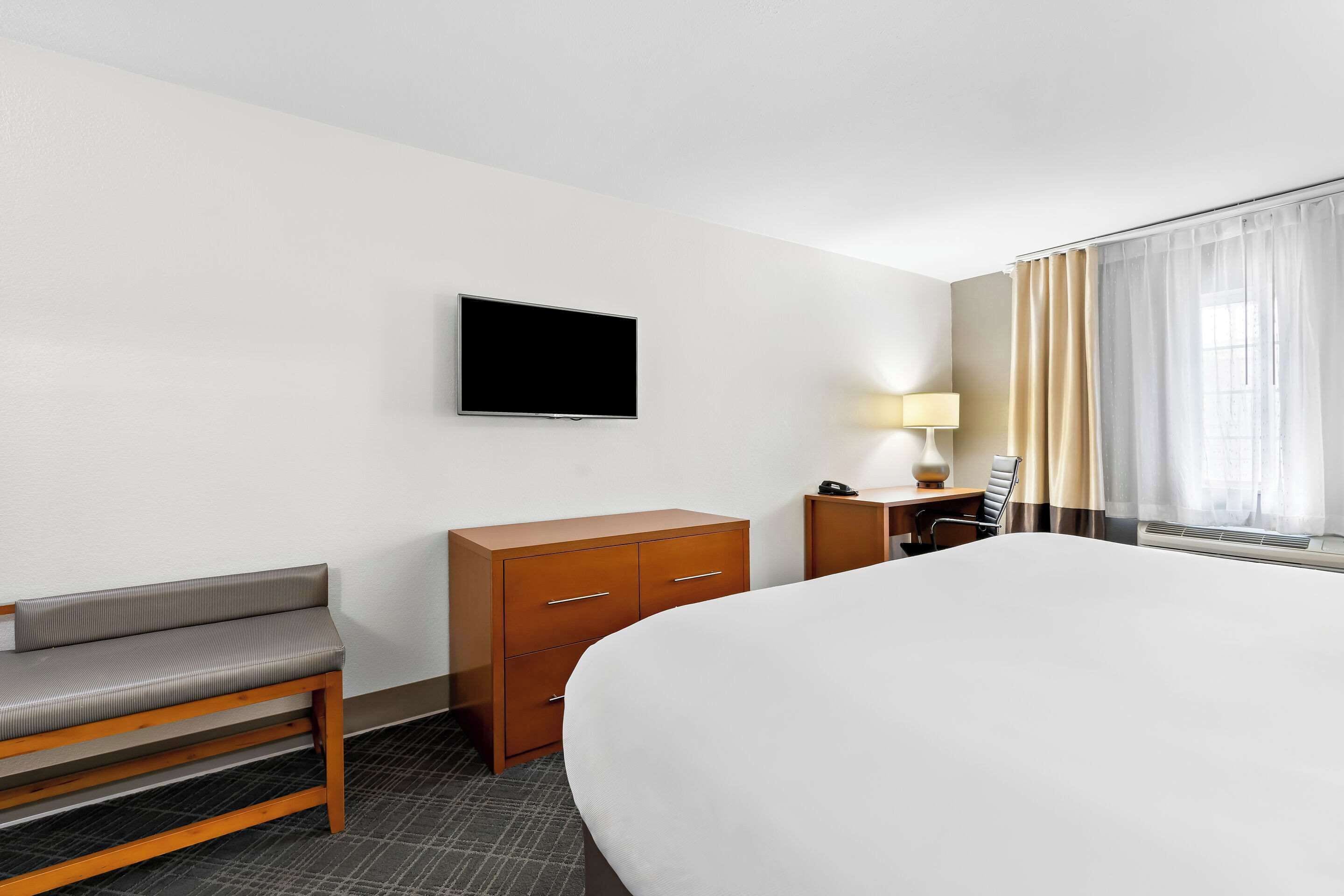 A hotel room with a large bed, a wooden bench, a desk with a lamp, a TV mounted on the wall, and a window with curtains.