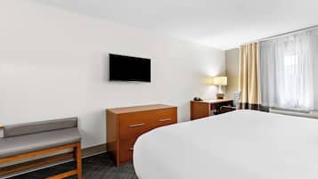 A hotel room with a large bed, a wooden bench, a desk with a lamp, a TV mounted on the wall, and a window with curtains.