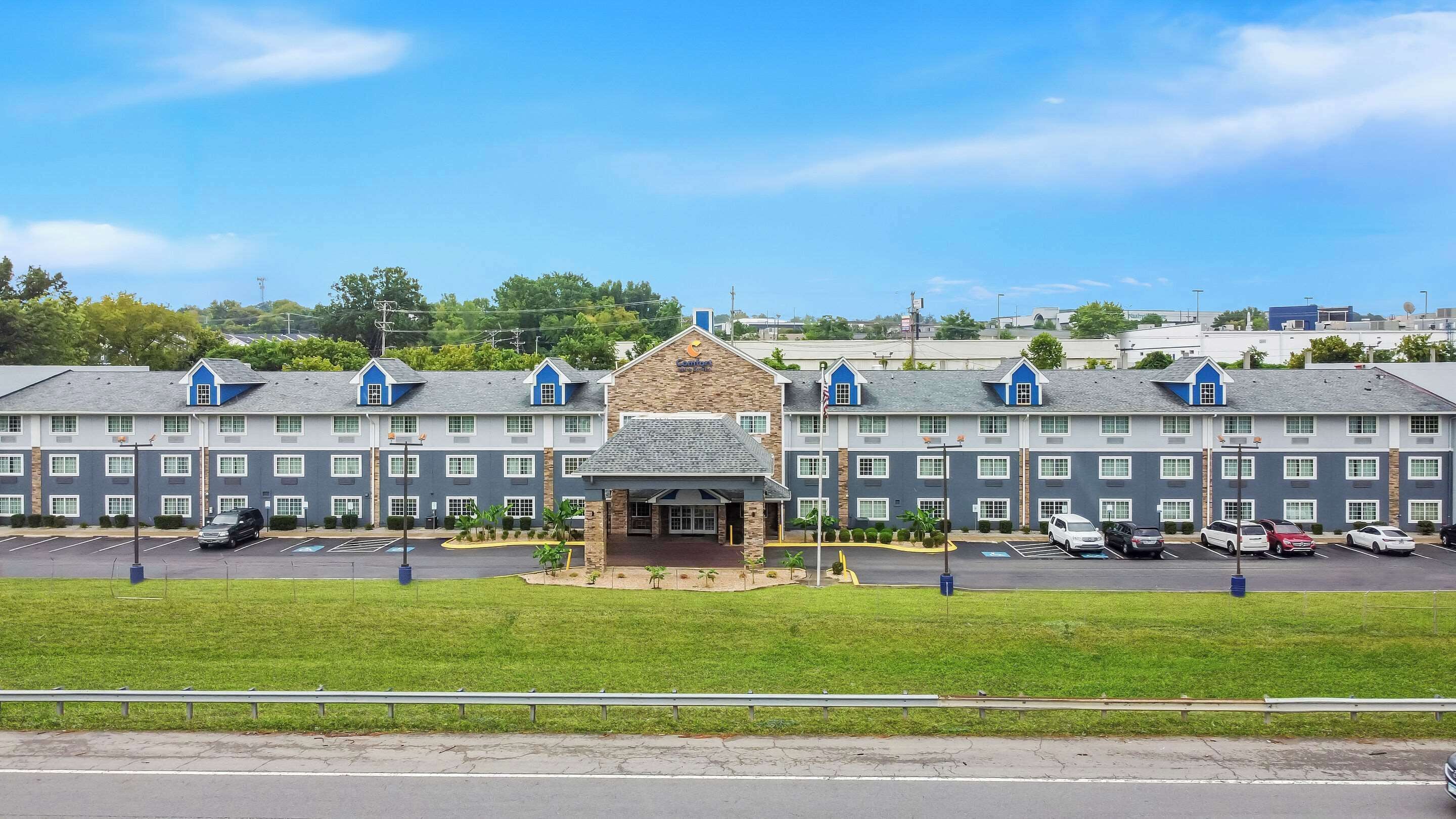 A multi-story hotel with a central entrance, parking lot, and a grassy area in front.