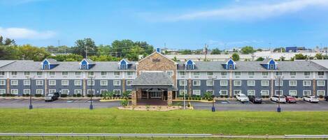 A multi-story hotel with a central entrance, parking lot, and a grassy area in front.