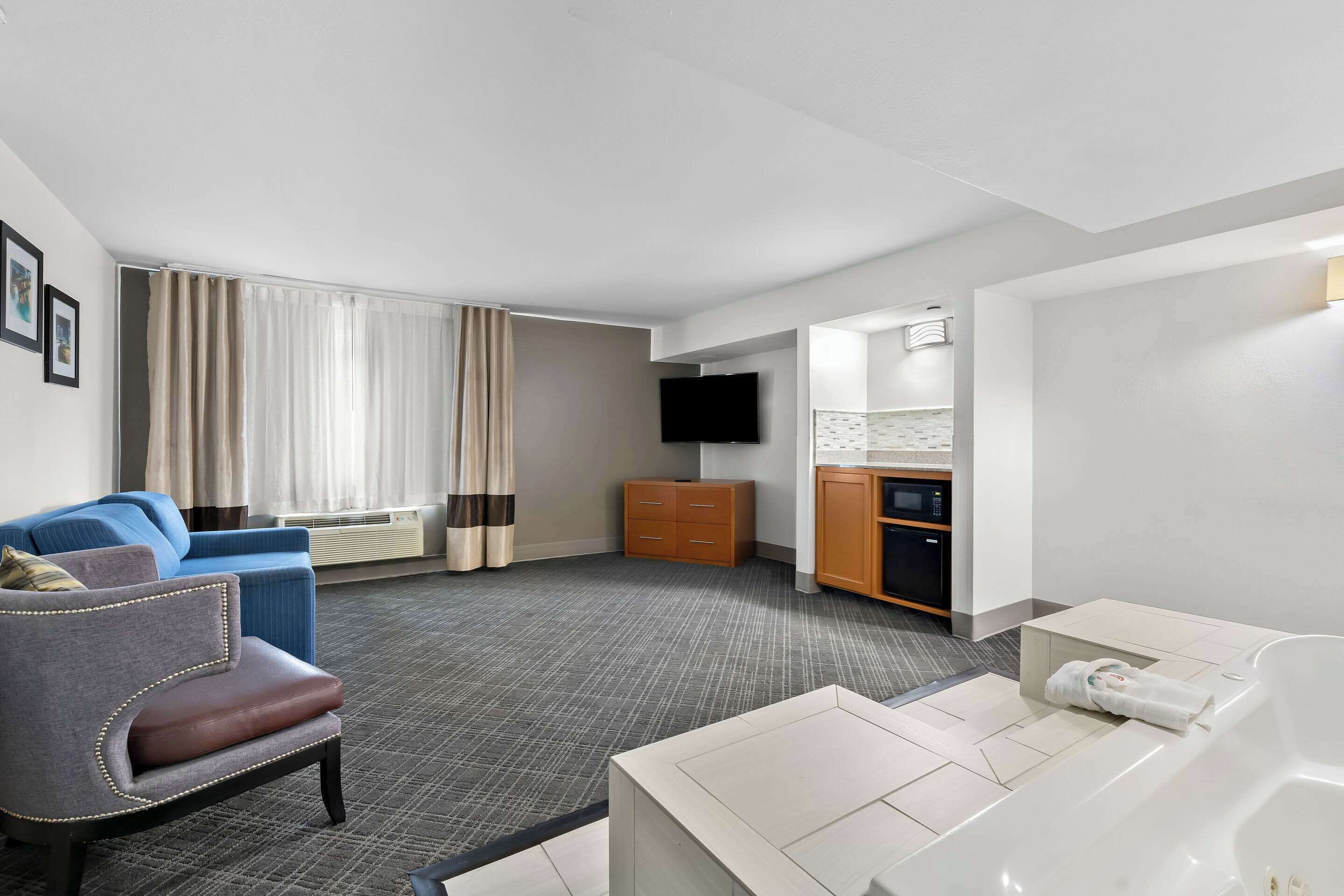Ein Hotelzimmer mit grauem Sessel, blauem Sofa, weißem Sofa, einem Flachbildfernseher, einem Holzschrank und einer kleinen Küchenzeile.