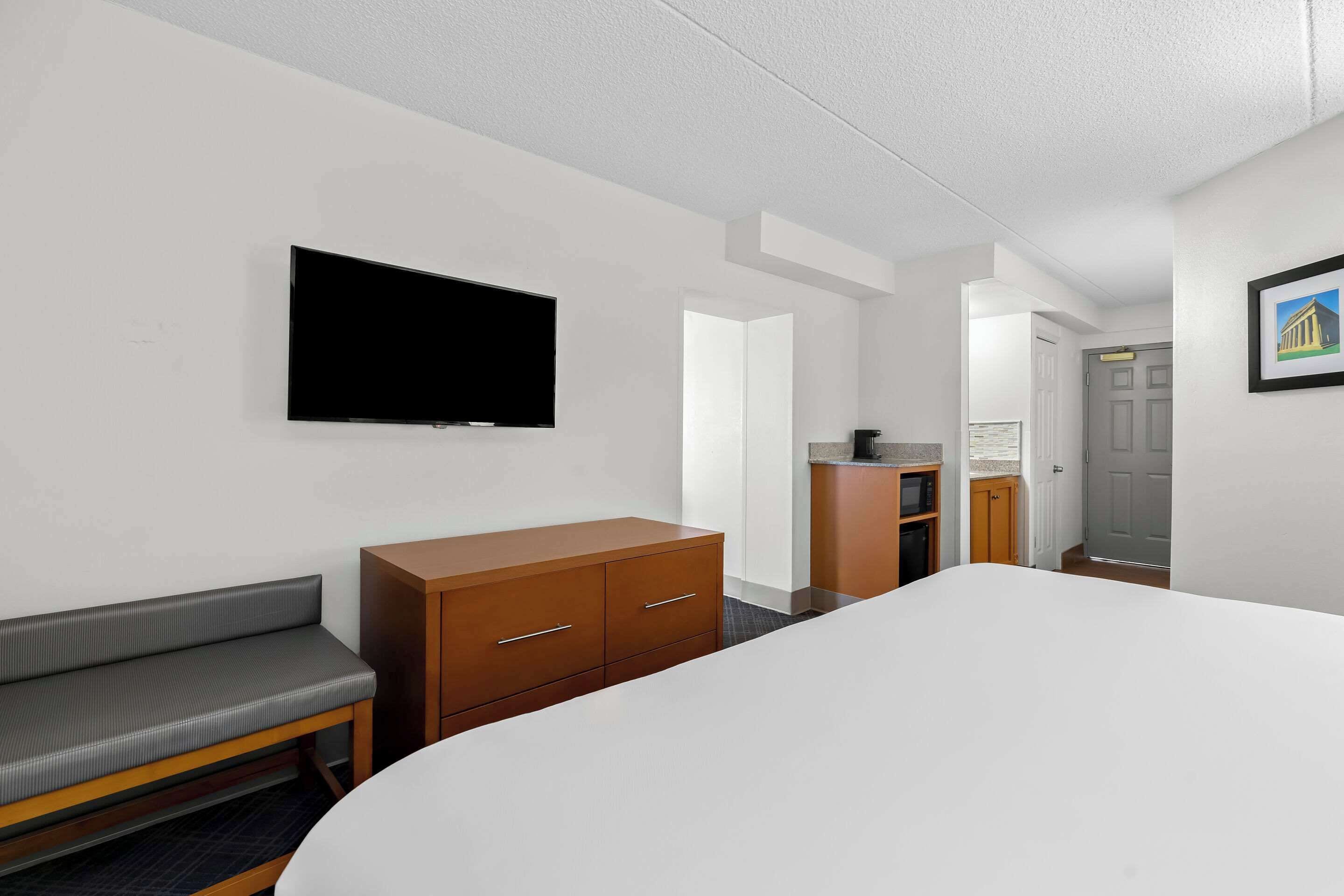 A hotel room with a bed, a TV mounted on the wall, a wooden dresser, a bench, and a door leading to another room.