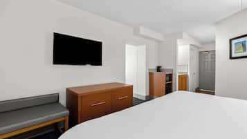 A hotel room with a bed, a TV mounted on the wall, a wooden dresser, a bench, and a door leading to another room.