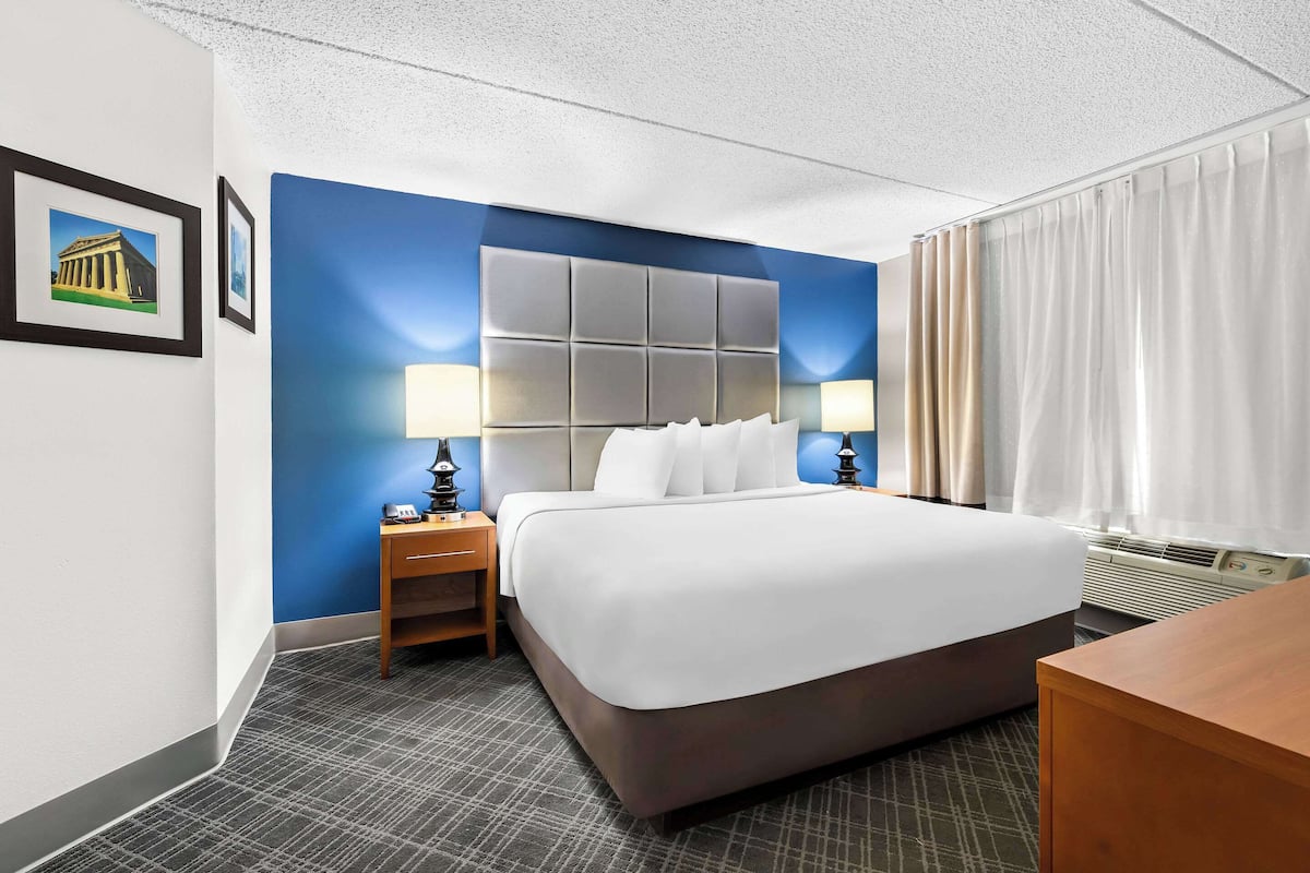 A hotel room with a large bed, two bedside tables with lamps, a small wooden nightstand, a wall-mounted air conditioning unit, and a window with curtains.