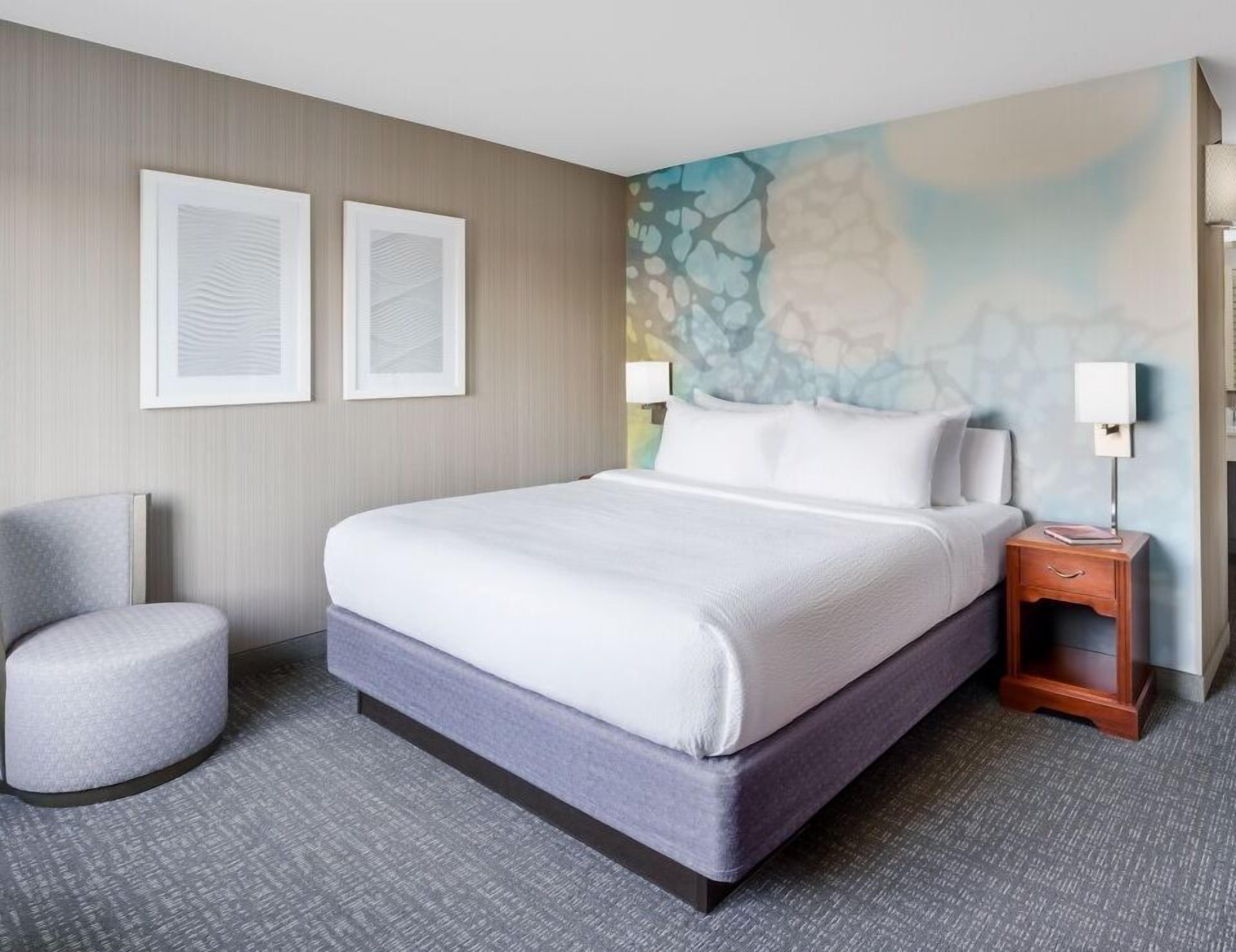 A hotel room with a bed, a chair, a nightstand, and a wall with abstract art.