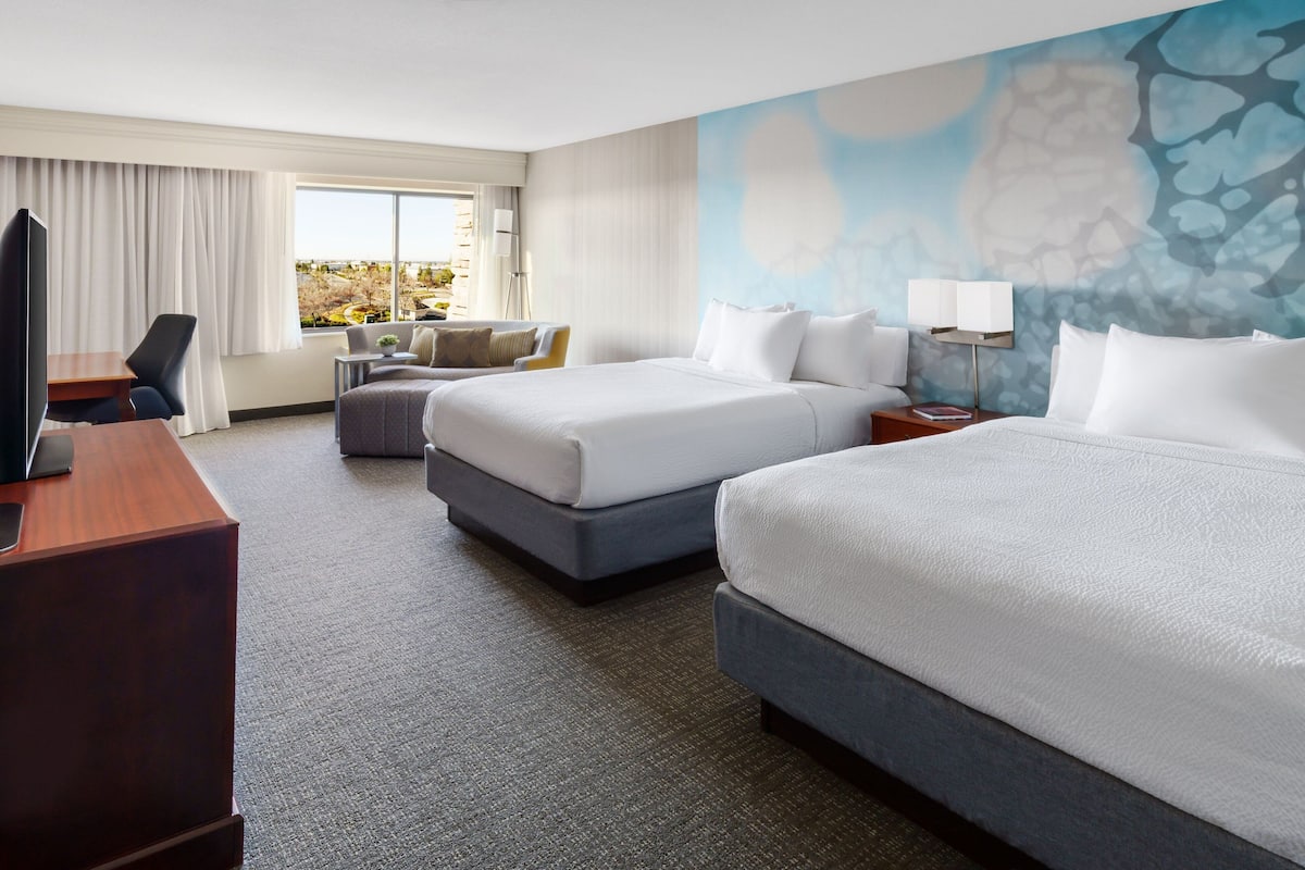 A hotel room with two beds, a desk, a chair, a TV, and a window with a view.
