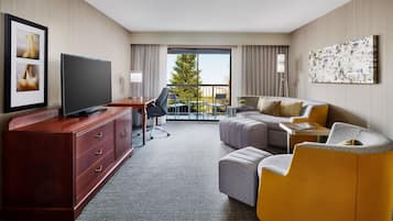 A modern hotel room with a flat-screen TV, a wooden dresser, a sofa, a chair, and a balcony with a view of trees.