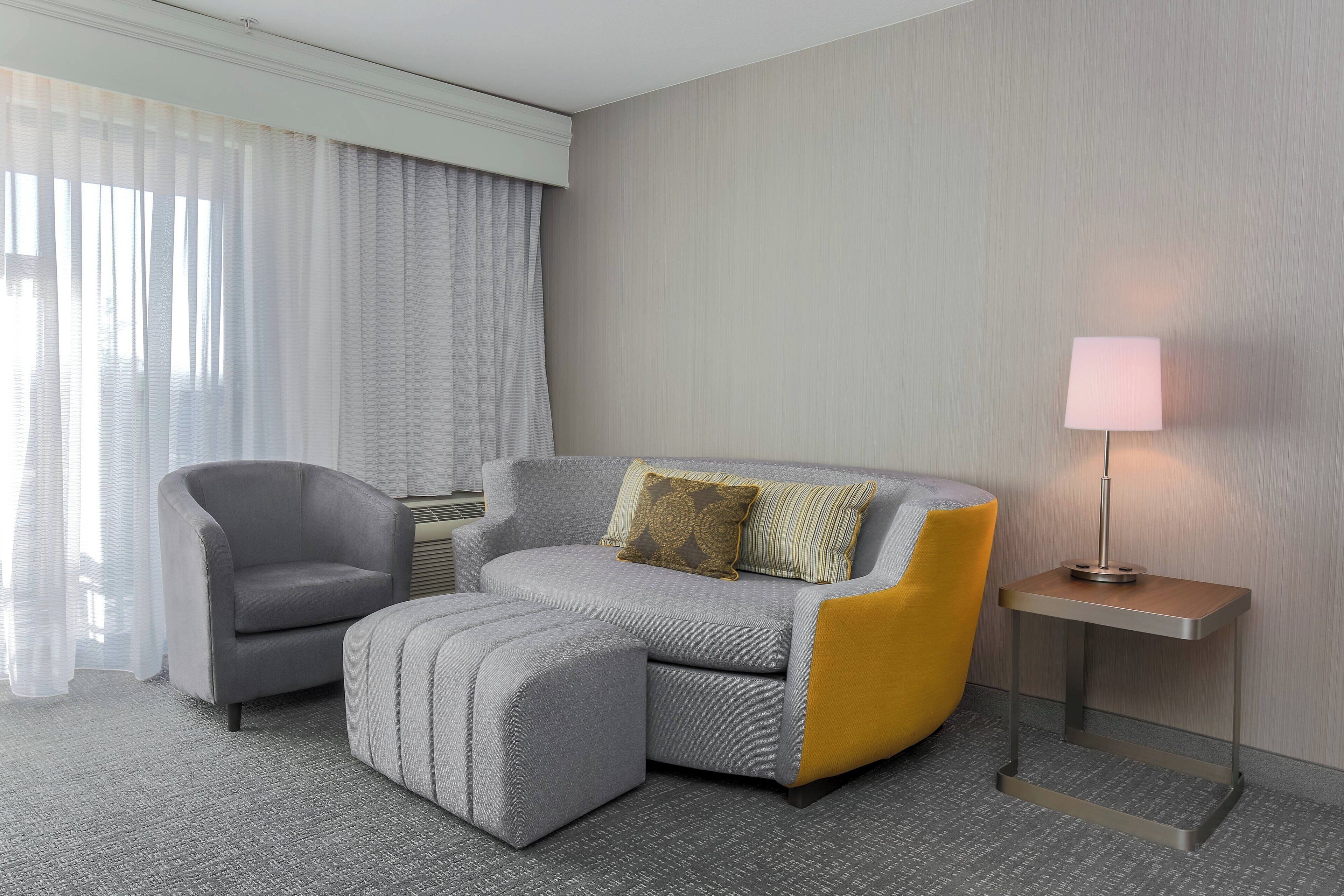 Moderni hotellihuone, jossa on harmaa sohva, samanvärinen jalkarahi, keltainen nojatuoli ja yöpöytä, jossa on lamppu.