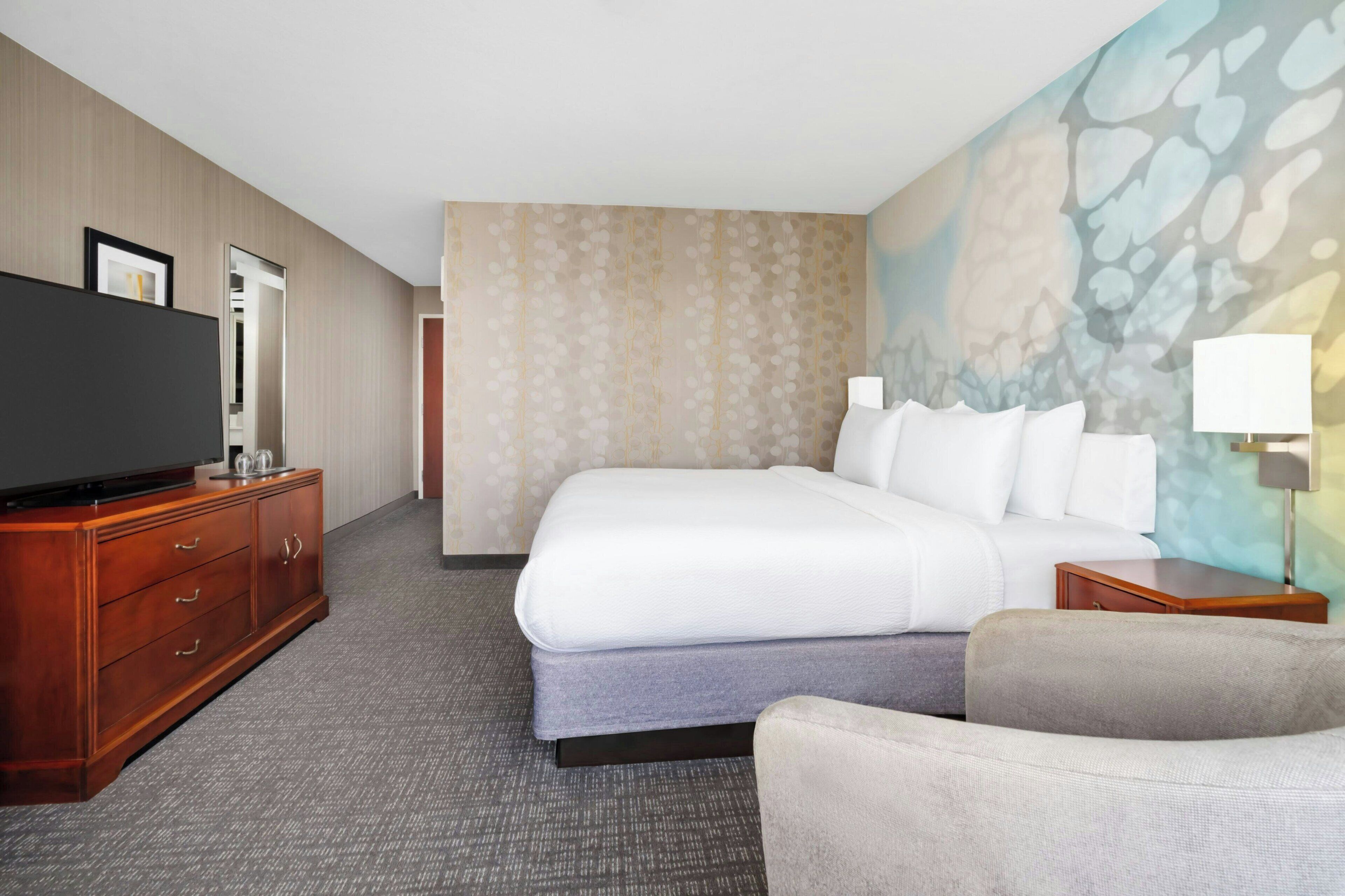 A hotel room with a large bed, a TV on a wooden dresser, a chair, and a painting on the wall.