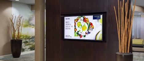 A reception area with a digital menu board advertising a farm stand breakfast bowl, a large potted plant, and a tall decorative vase.